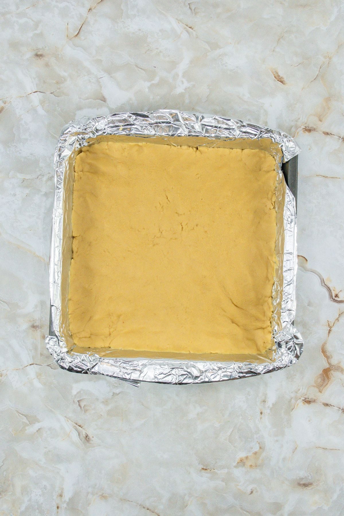 a pan lined with foil, containing a layer of creamy peanut butter, unsalted butter, and powdered sugar mixture
