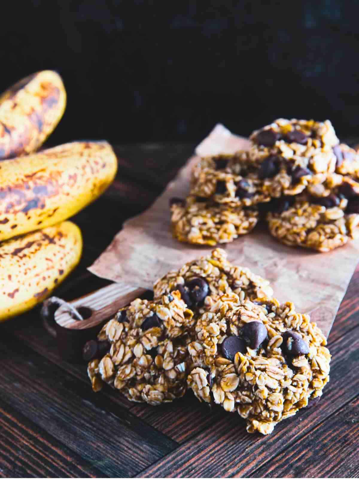 A pile of banana oatmeal cookies on a cutting board with ripe bananas in the background.