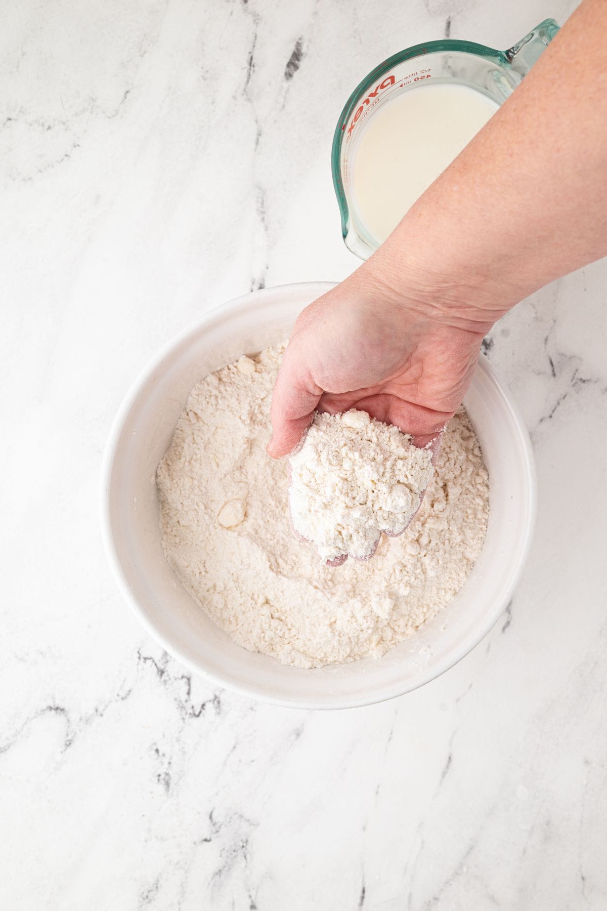 A hand holding flour mixture while pouring milk into a bowl.