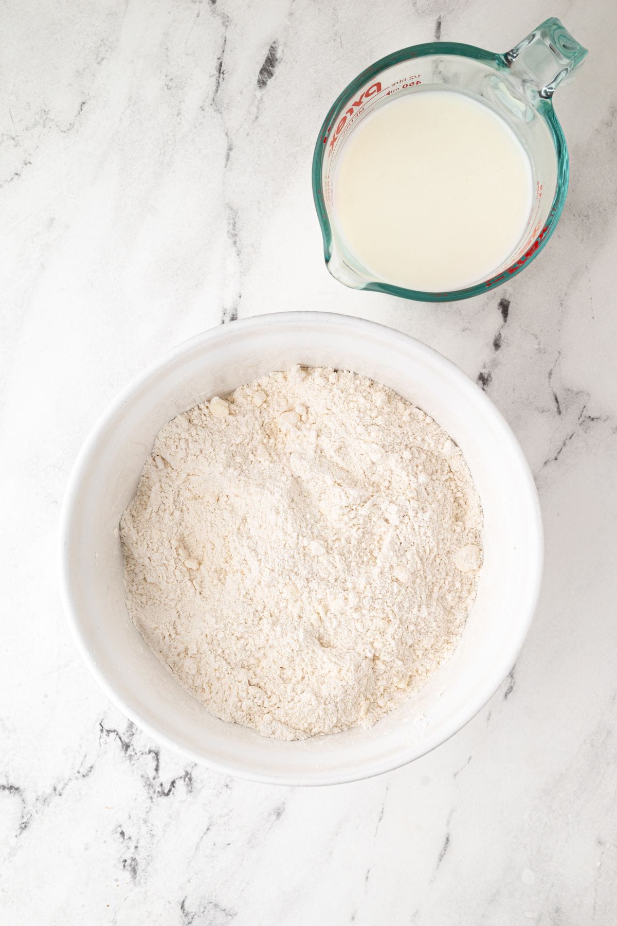A bowl with dry ingredients next to a measuring cup filled with milk.