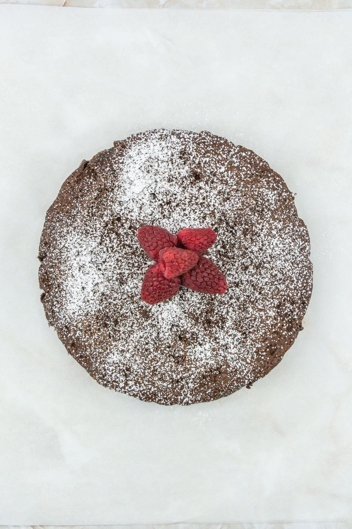 a round chocolate cake dusted with powdered sugar, topped with a cluster of fresh raspberries arranged in the center