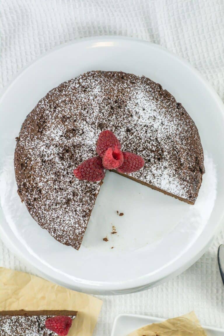 overhead shot of a round chocolate cake with a slice taken out, dusted with powdered sugar, and topped with fresh raspberries