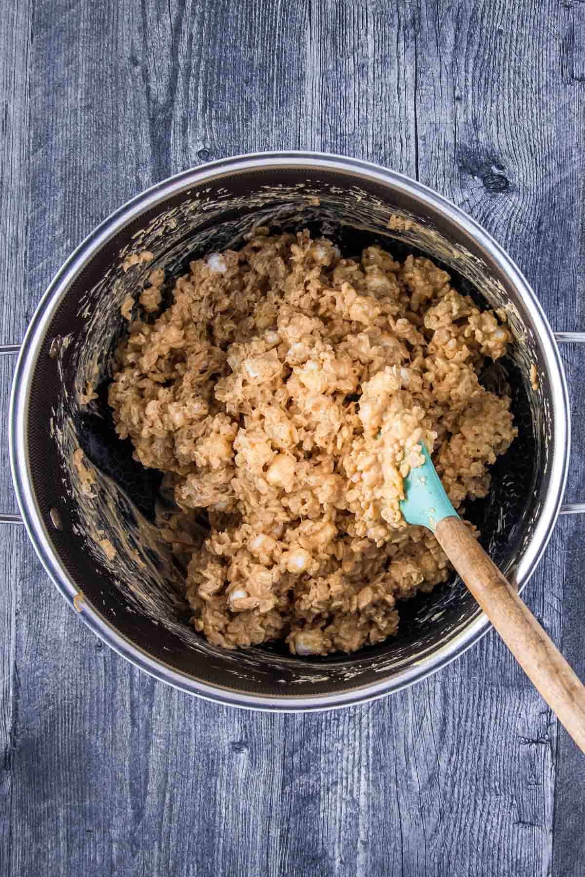 melted mixture of white chocolate chips and creamy peanut butter, mini marshmallows, and rice krispies cereal being mixed by a spatula in a large pot