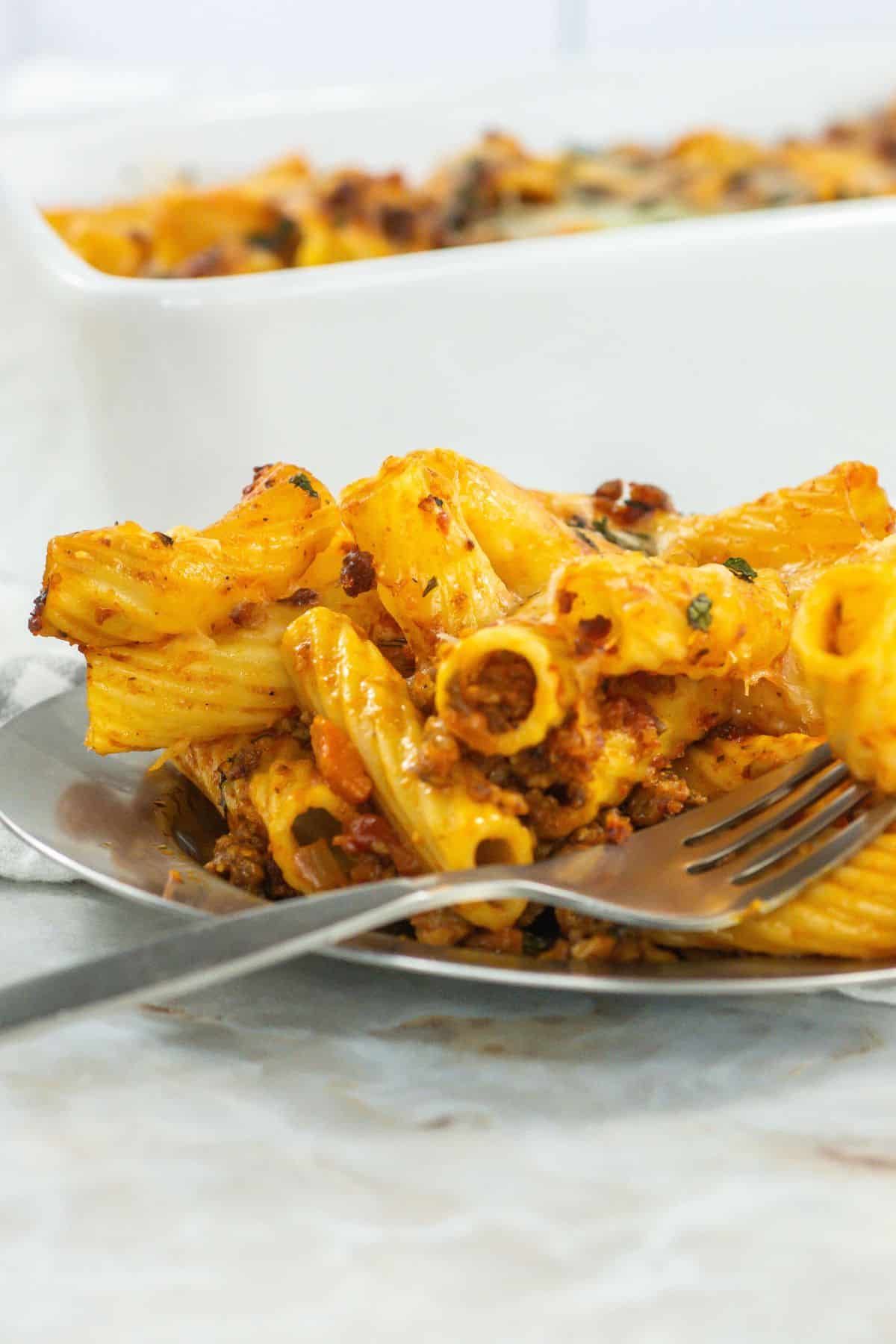 Close-up of plated serving: A fork resting on a serving of saucy baked rigatoni topped with melted cheese and herbs.