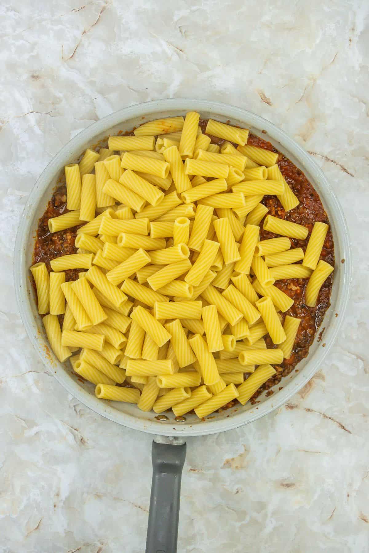 Cooked rigatoni being stirred into the meat sauce in a skillet, evenly coated.
