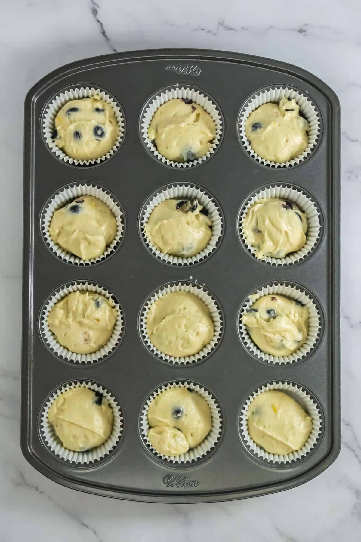 Cupcake batter portioned into liners in a muffin tin, some with visible blueberries.