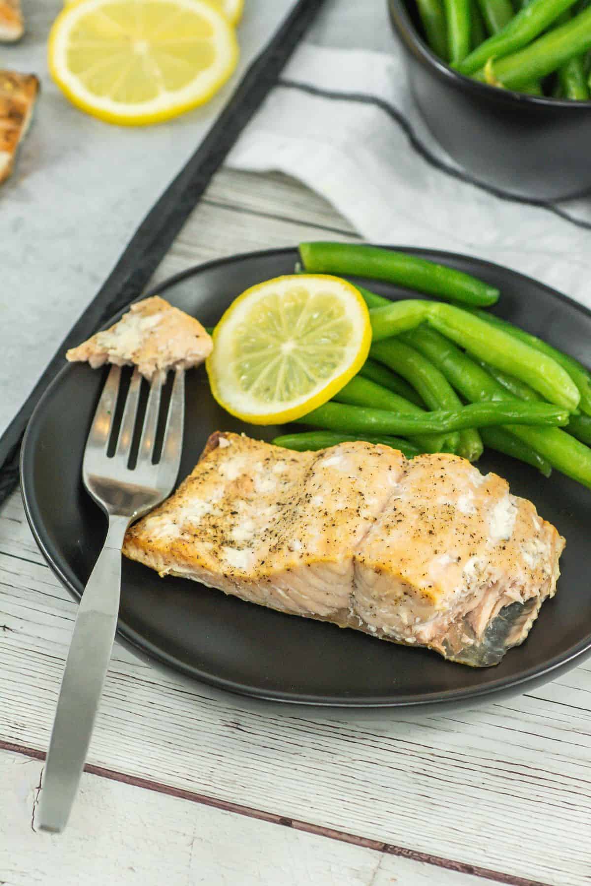 A broader view of the cooked salmon plated with a fork cutting through the flaky fish. The serving also includes green beans and lemon wedges on a dark plate, highlighting a complete meal presentation.