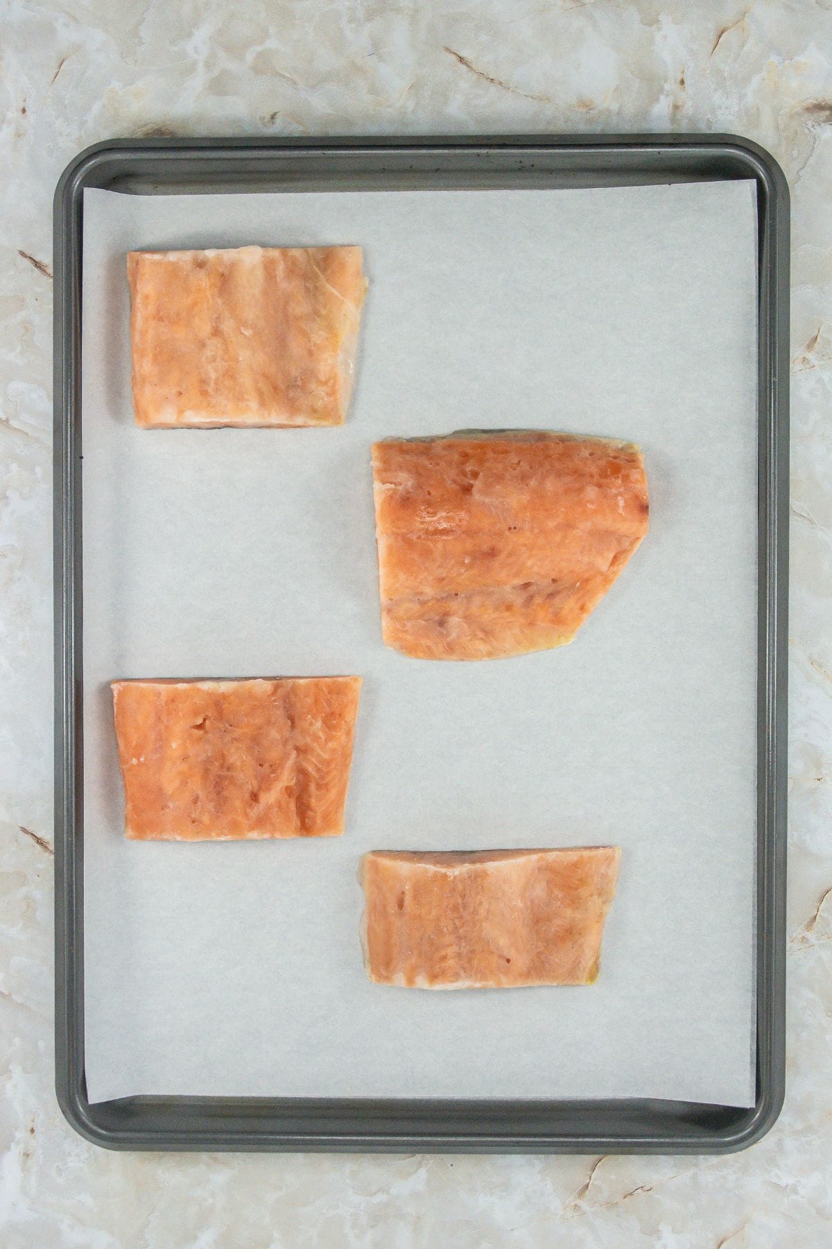 This image shows raw salmon fillets arranged on a baking sheet lined with parchment paper. The fillets appear frozen, with a slight sheen from ice crystals or moisture. The focus is on the preparation phase before seasoning or cooking.