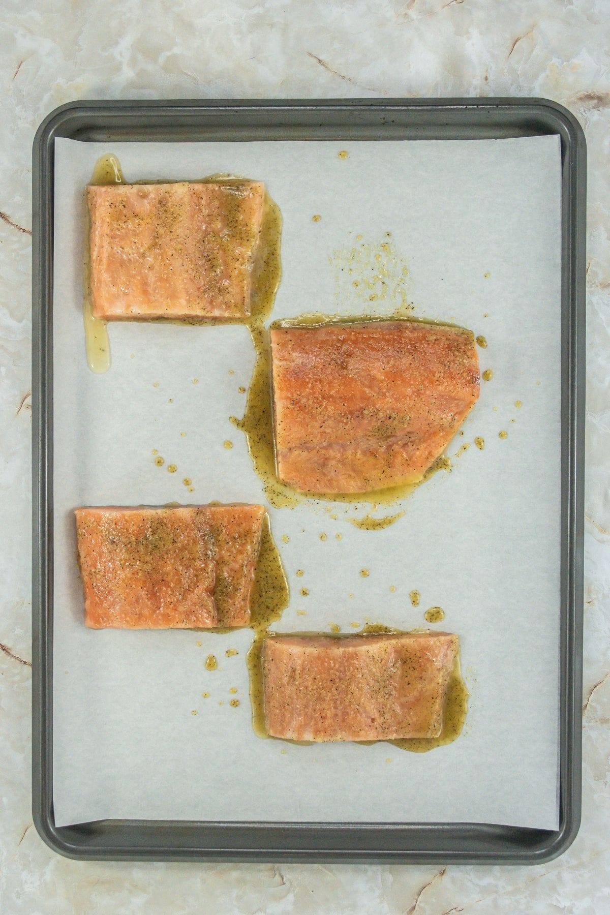 The salmon fillets on a parchment-lined baking sheet have been seasoned with a liquid marinade or oil-based dressing. The seasoning creates a glossy layer on the surface, indicating they are ready for broiling.