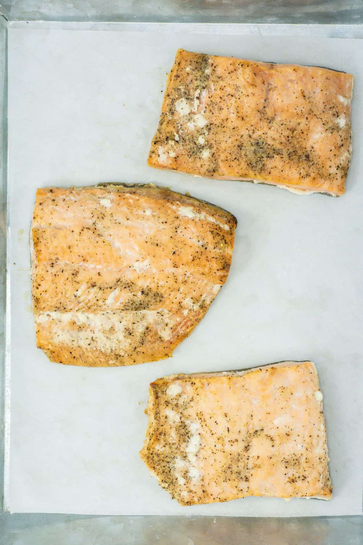 Three broiled salmon fillets on parchment paper, seasoned and fully cooked.