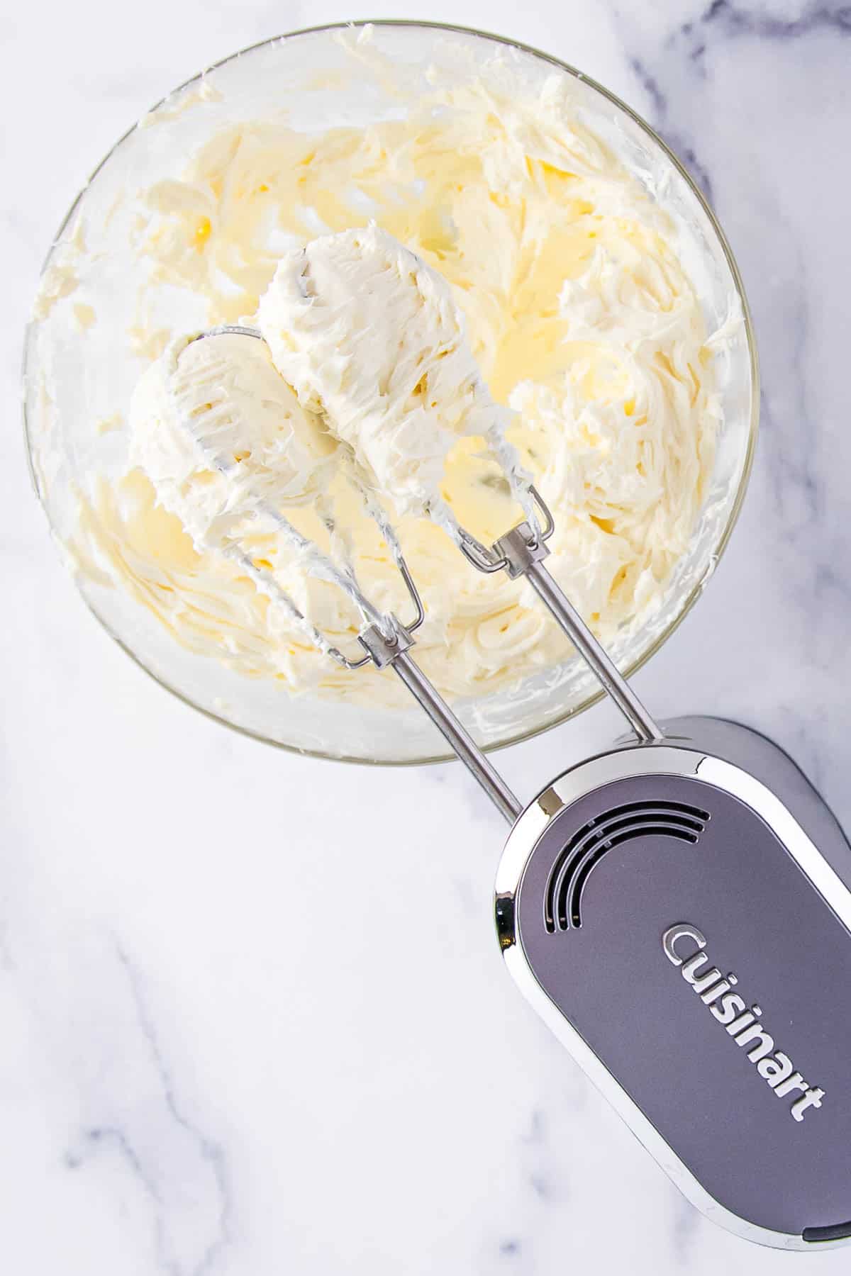 an electric mixer with beaters covered in cream cheese on top of a bowl containing more cream cheese