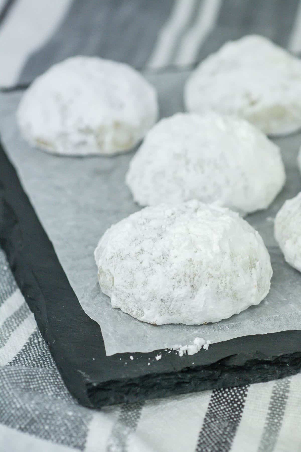 A slate black tray lined with parchment paper holds six Butter Pecan Meltaways. The cookies are neatly arranged, showing their snow-white powdered sugar coating and soft, dome