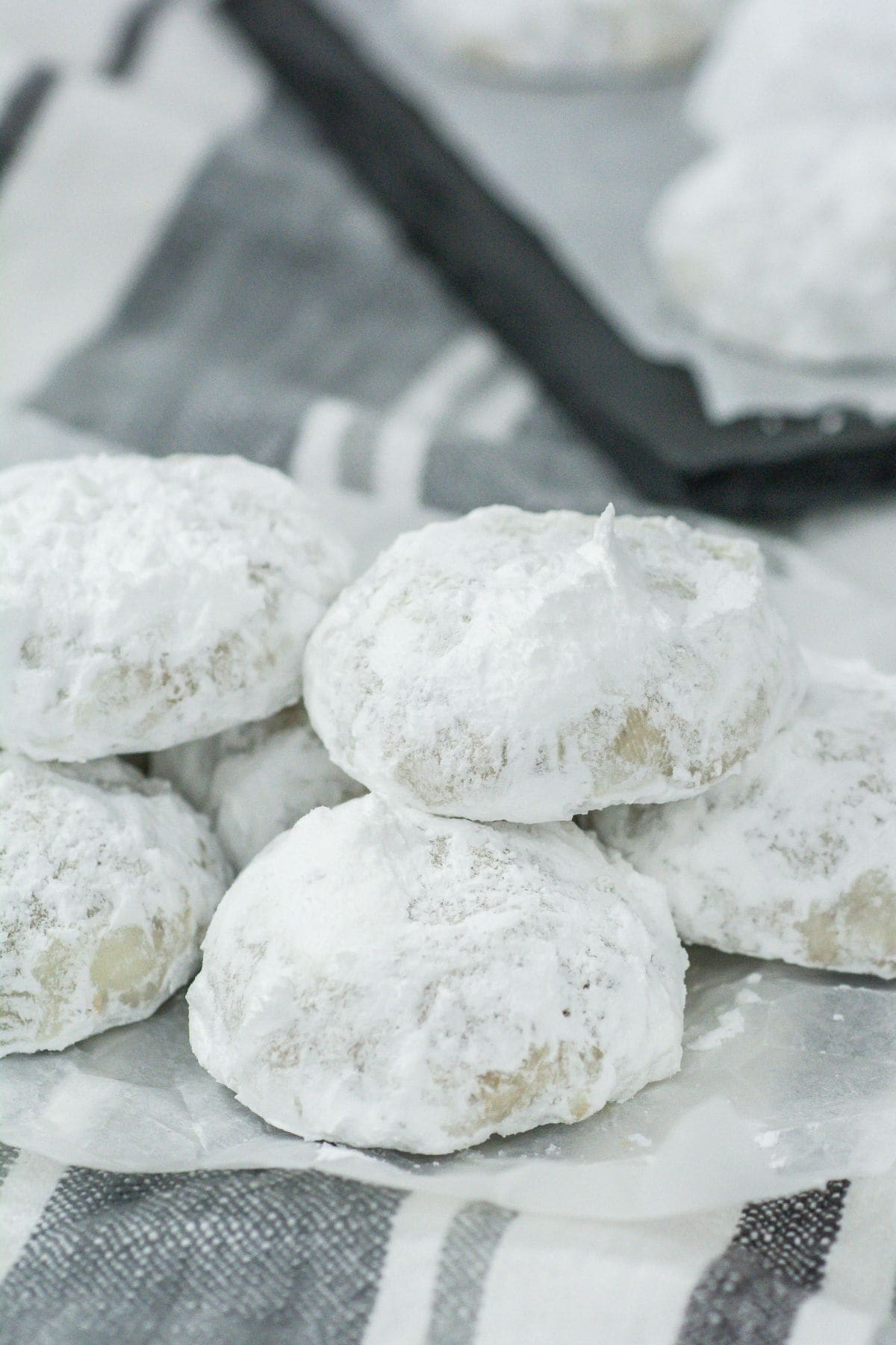A close-up of a pile of finished snowball cookies. The powdered sugar generously coats each cookie, highlighting their soft, rounded shapes. They are resting on parchment paper with a striped cloth background.