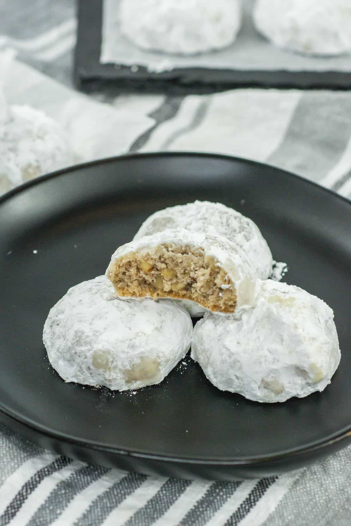A close-up of three cookies on a black plate, with one cookie broken in half to highlight the inside texture. The visible chopped pecans and crumbly cookie interior contrast beautifully with the powdered sugar coating.