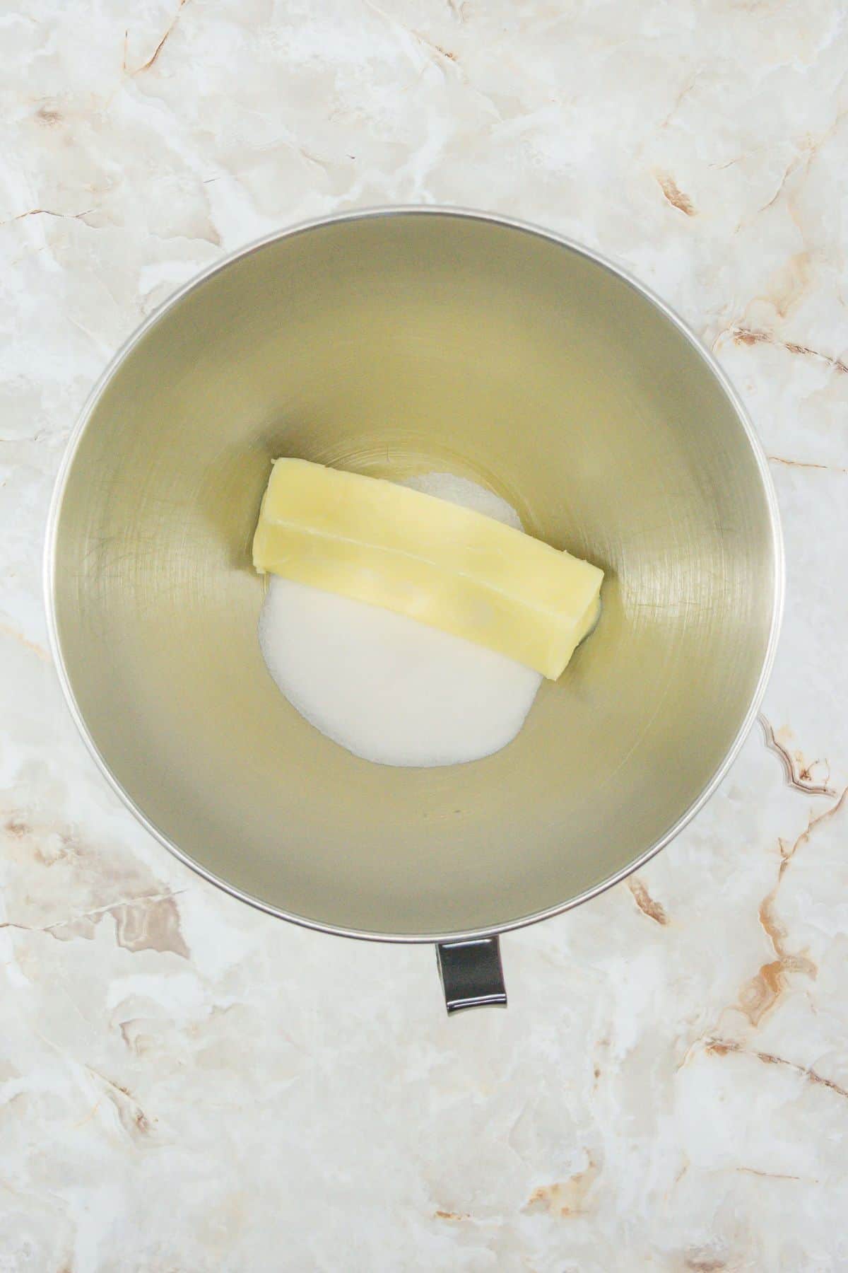 A stainless-steel bowl holding two ingredients: a stick of butter and a mound of white granulated sugar. This is the starting step for creaming butter and sugar together.