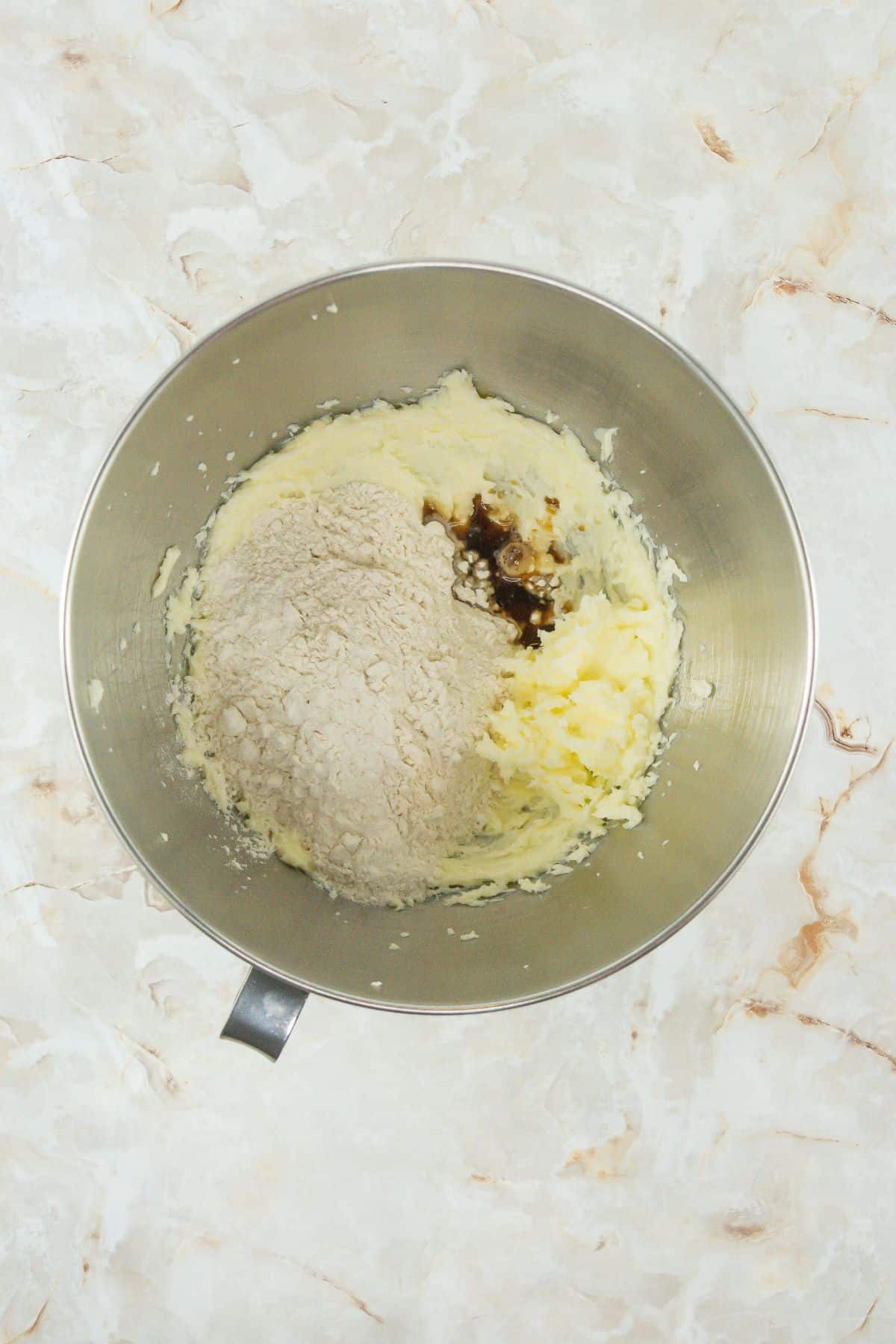 A mixing bowl showing the creamed butter and sugar mixture with flour and vanilla extract added but not yet mixed. This stage appears fluffy but powdery before combining.