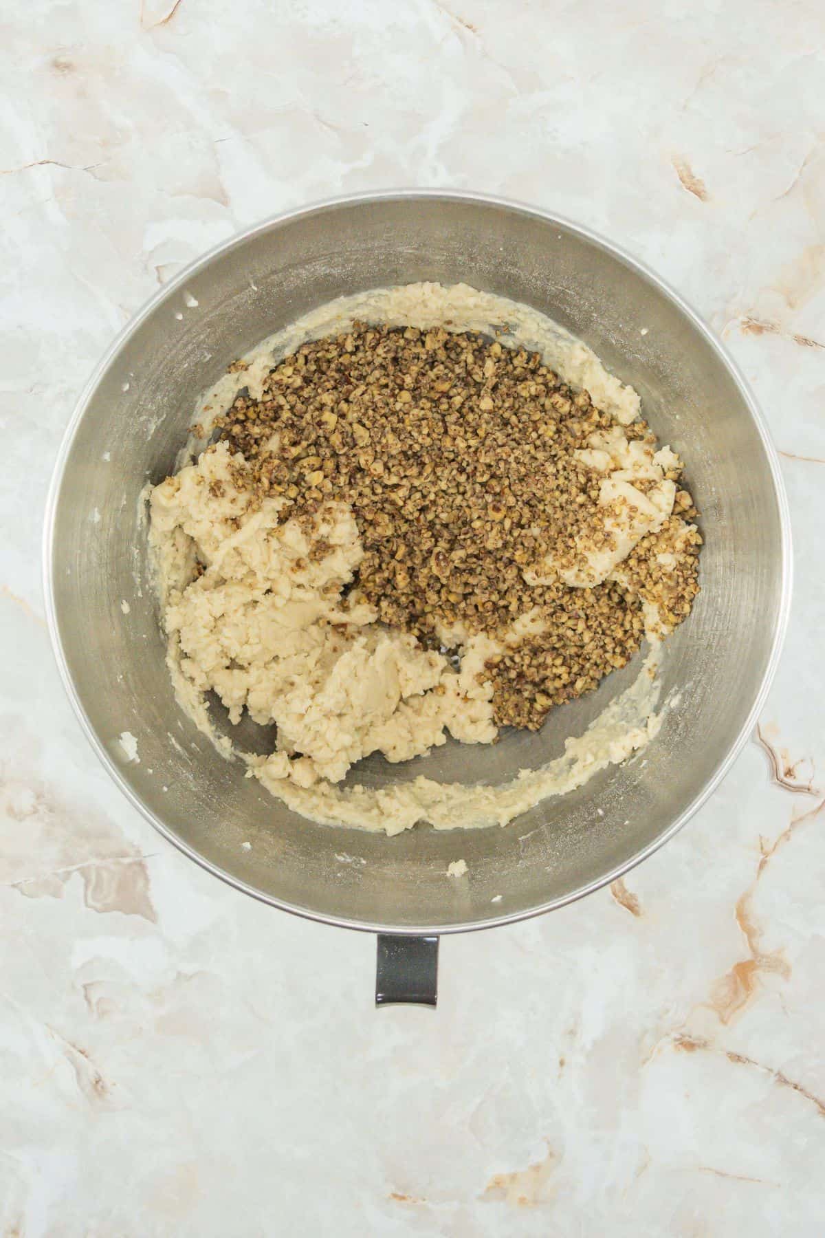 A large stainless-steel mixing bowl containing dough with a large portion of toasted, chopped pecans added on top. The pecans are evenly spread, about to be mixed into the dough.
