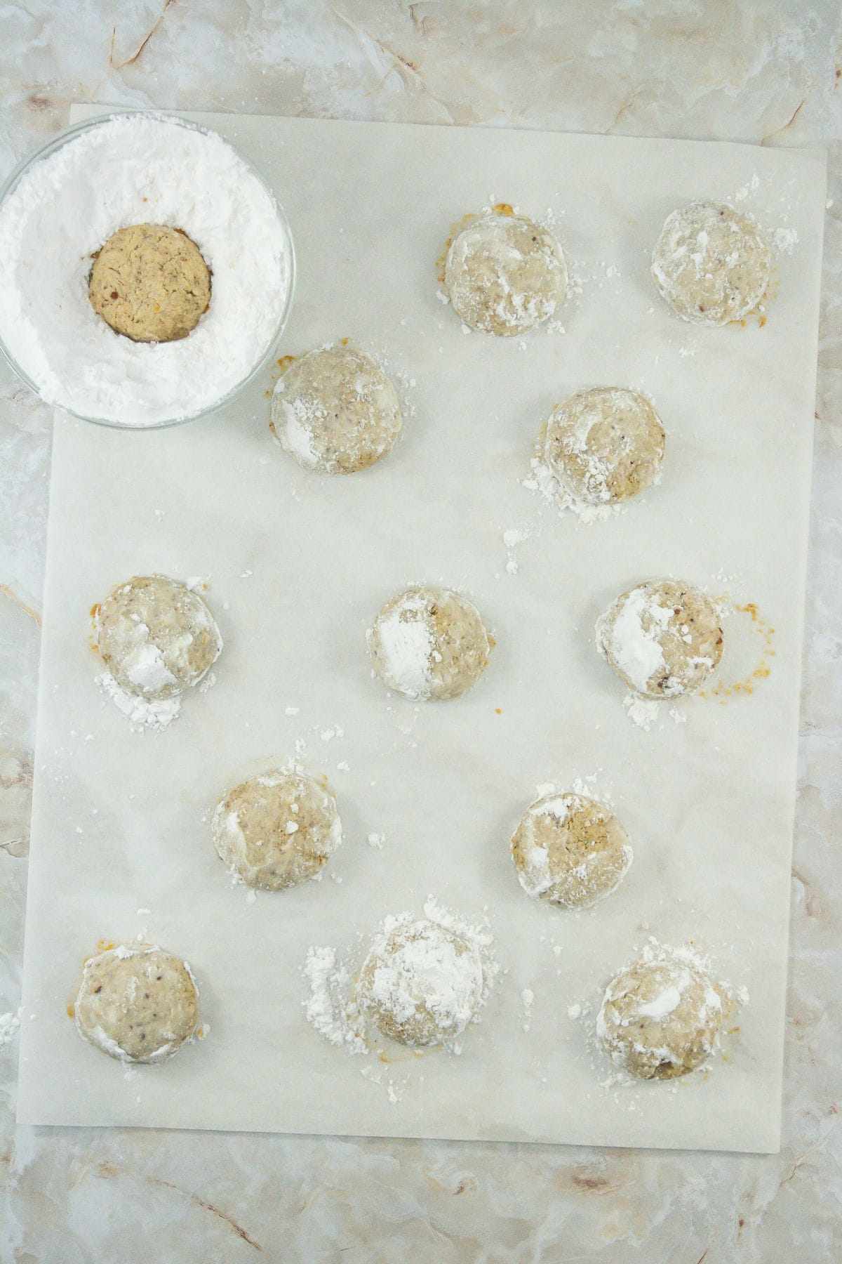 Rolled balls of cookie dough on parchment paper. One cookie ball is placed in a small bowl filled with powdered sugar, indicating the process of coating the cookies before baking.