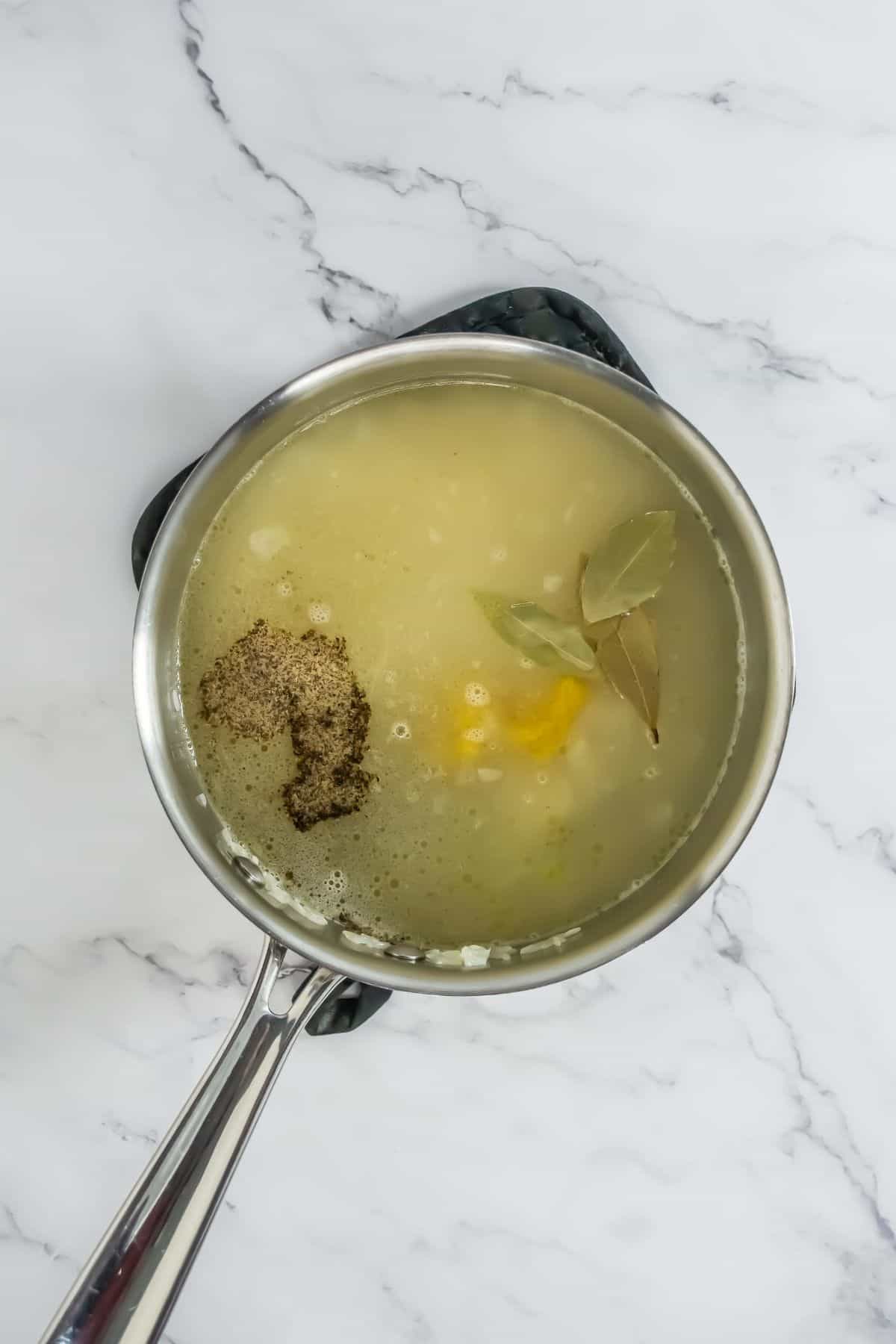 Broth, bay leaves, and seasoning added to the butter rice mixture in the saucepan, simmering.