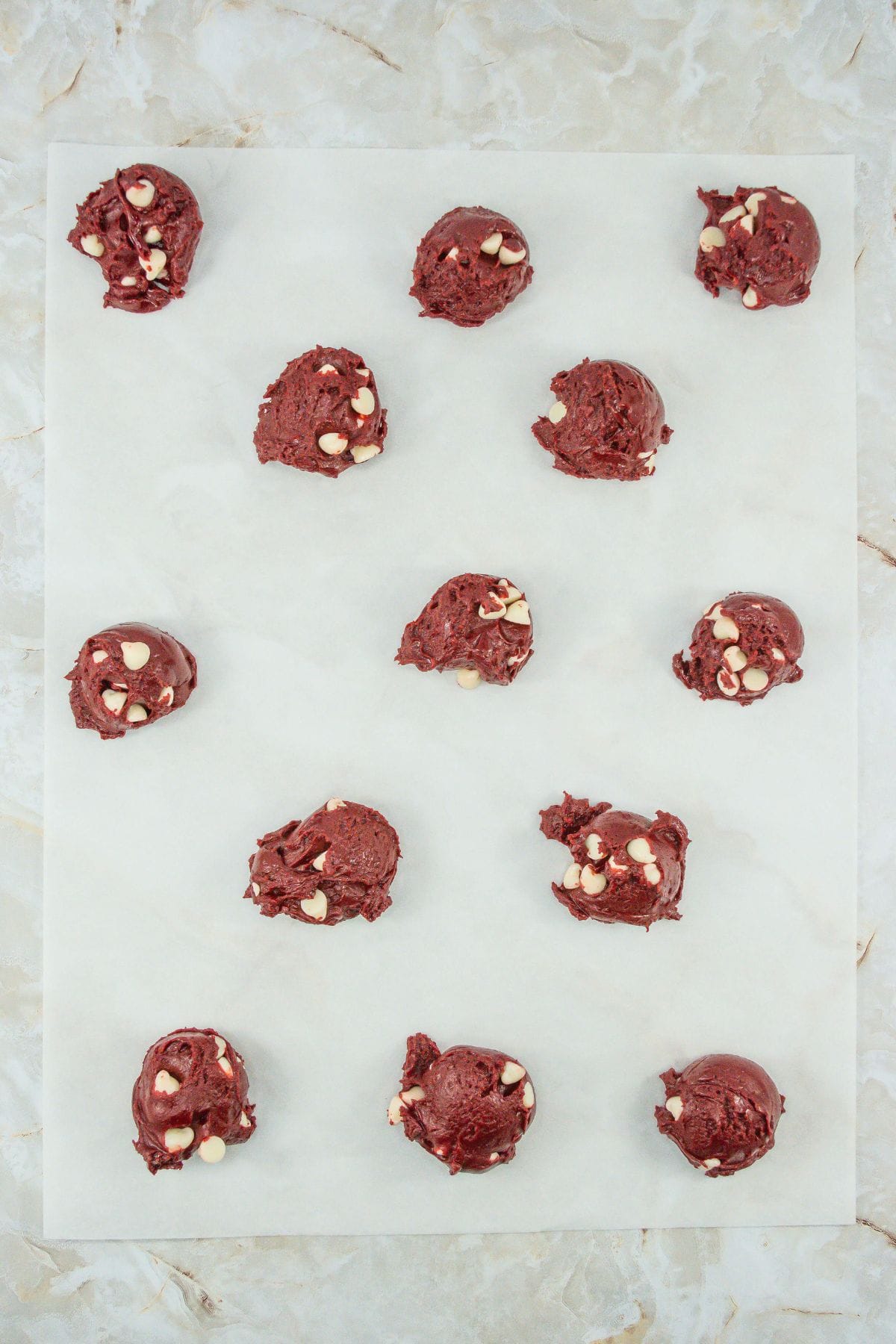 A parchment-lined baking tray with evenly spaced scoops of red velvet cookie dough mixed with white chocolate chips, ready to go into the oven.
