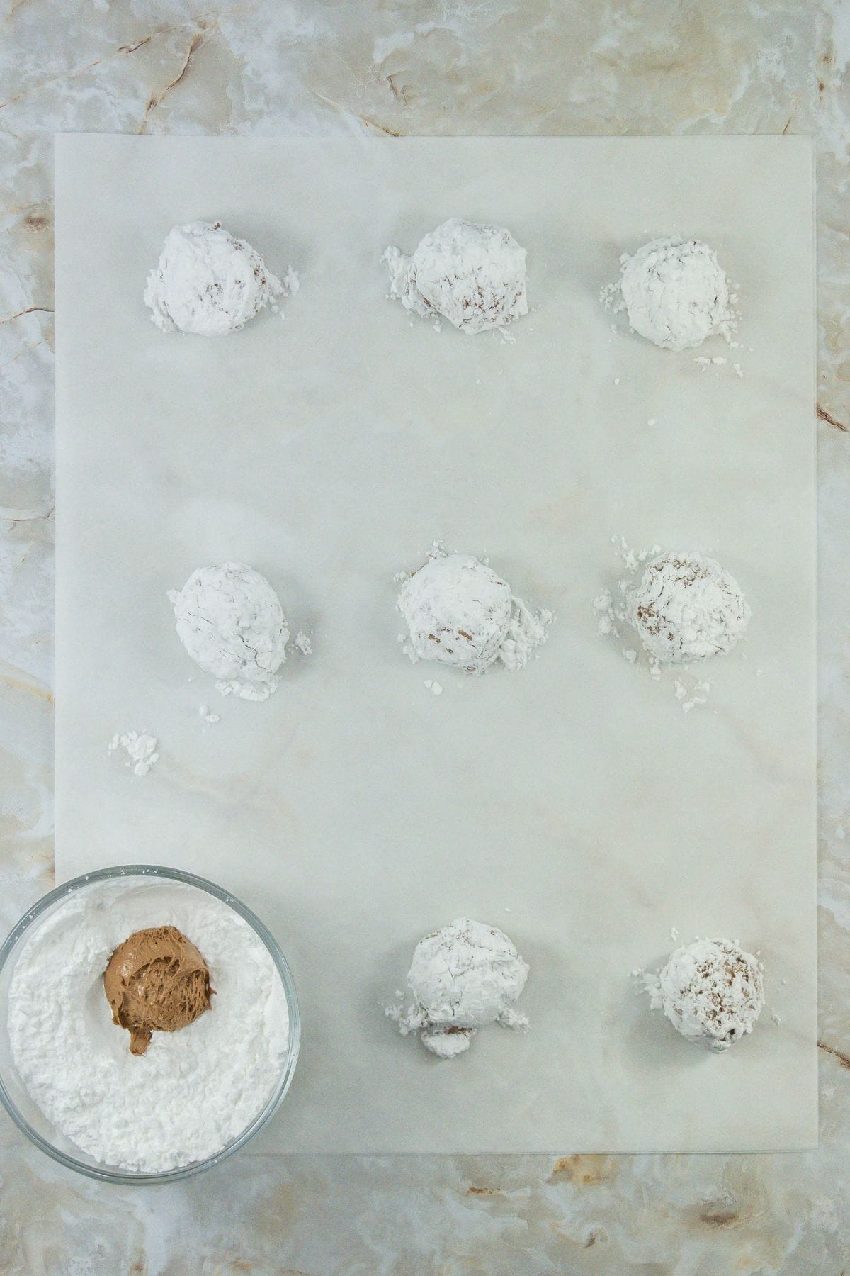 A parchment-lined surface with several cake mix and cool whip cookie dough balls rolled in powdered sugar, ready for baking.