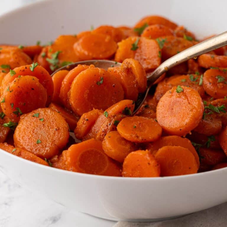 A large serving bowl filled with glossy, well-seasoned canned carrot slices, garnished with herbs and a spoon for serving.