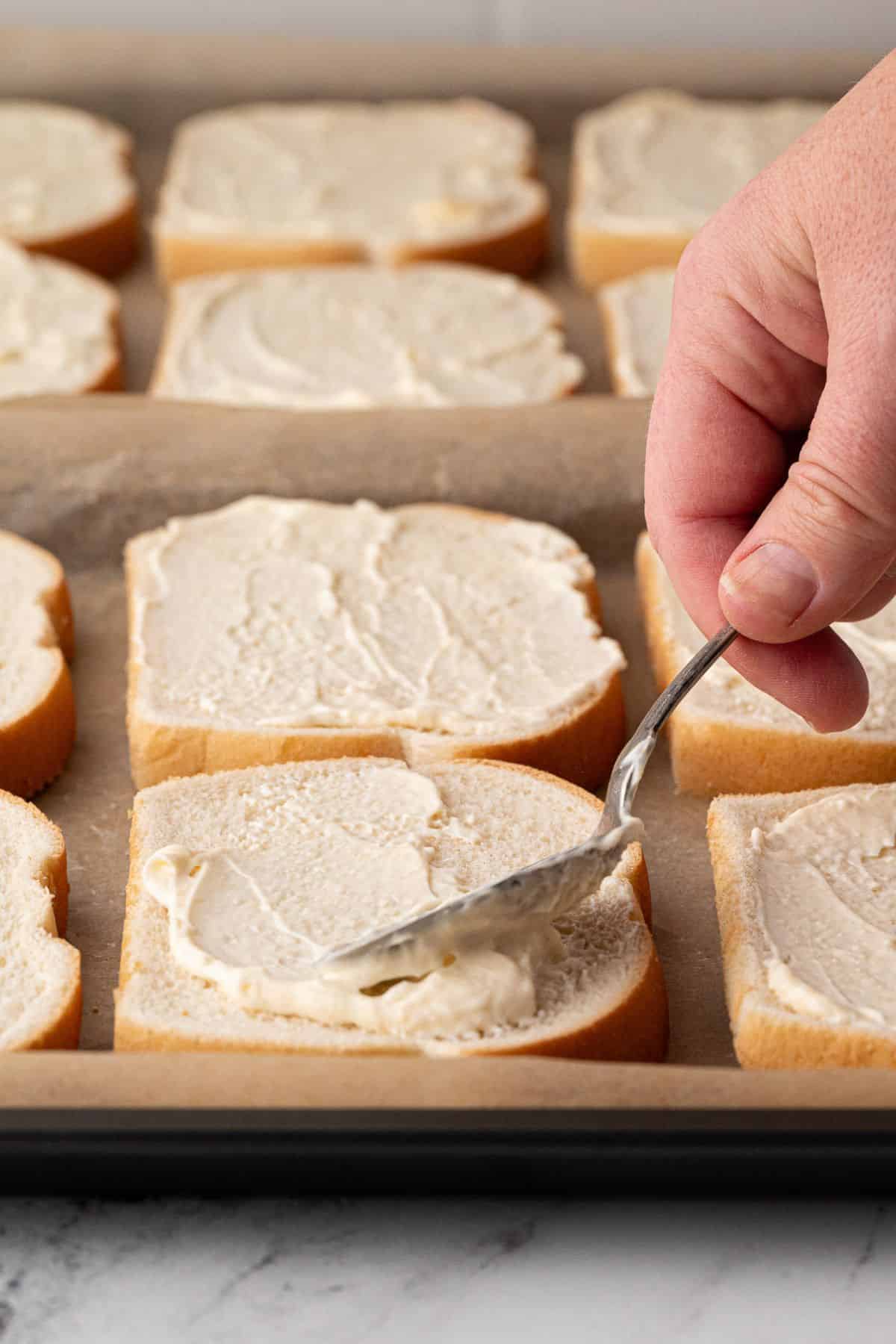 Hand spreading the creamy mixture onto sliced bread on a baking sheet lined with parchment.