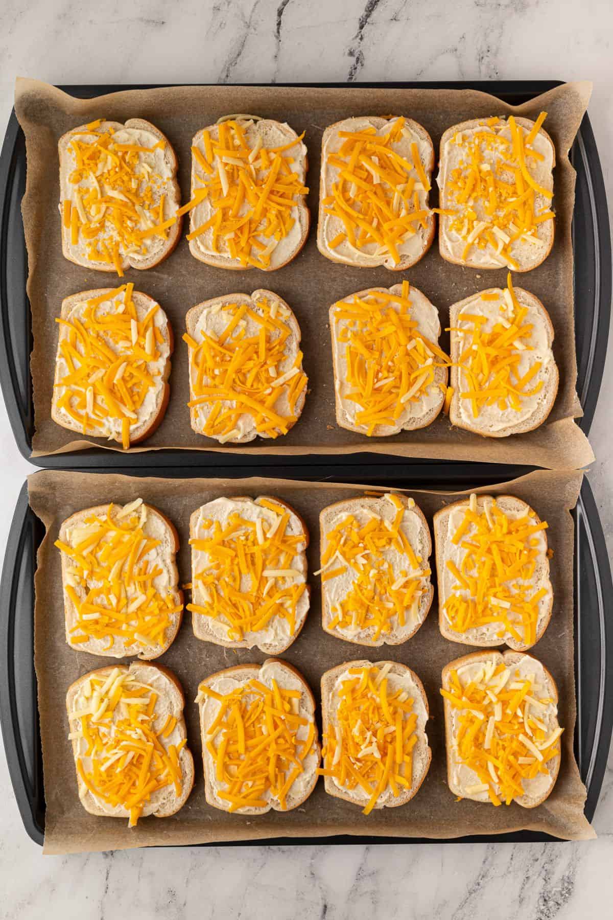 Two baking trays with multiple slices of bread topped with shredded cheese.