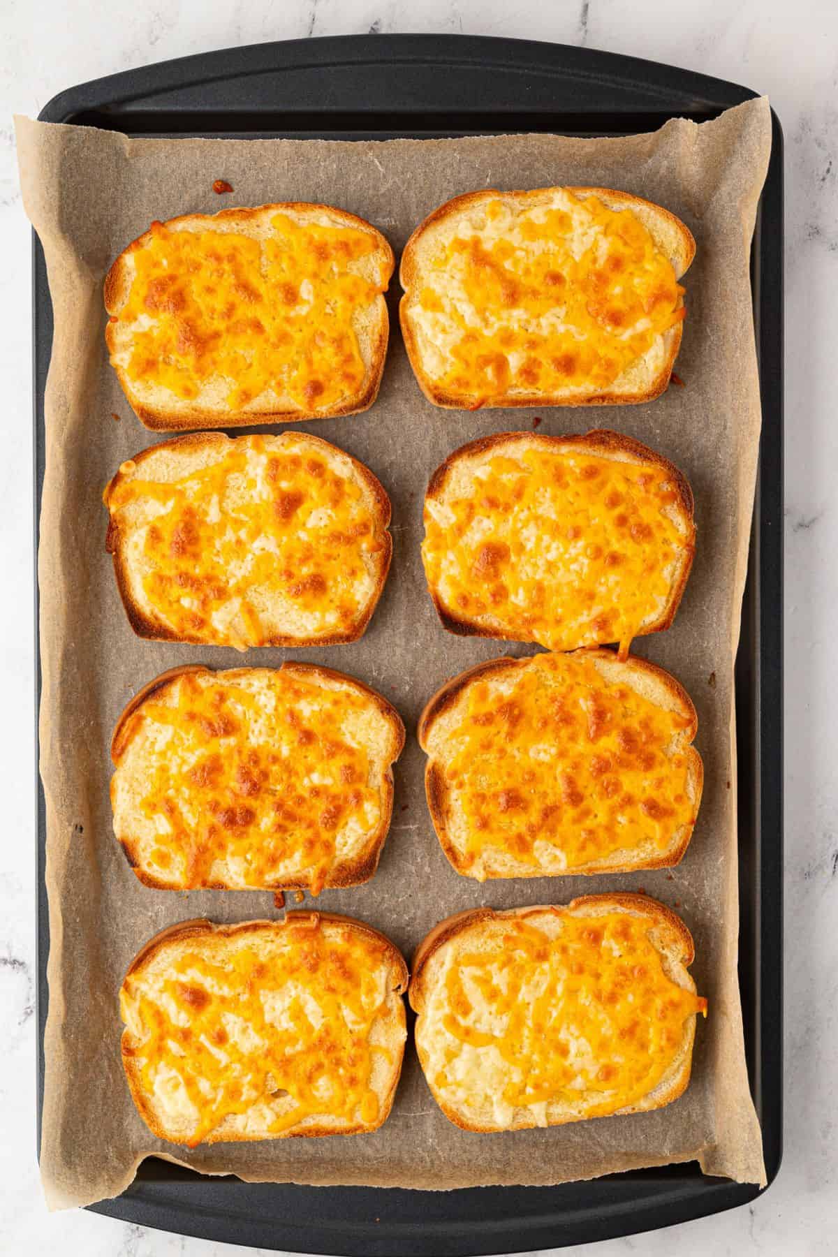 Eight perfectly baked cheese toasts arranged on a baking tray lined with parchment paper.