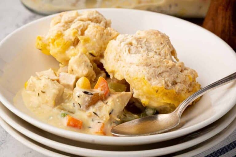 Bowl of creamy chicken pot pie casserole topped with golden brown biscuit dough, served with a spoon and stacked white plates underneath