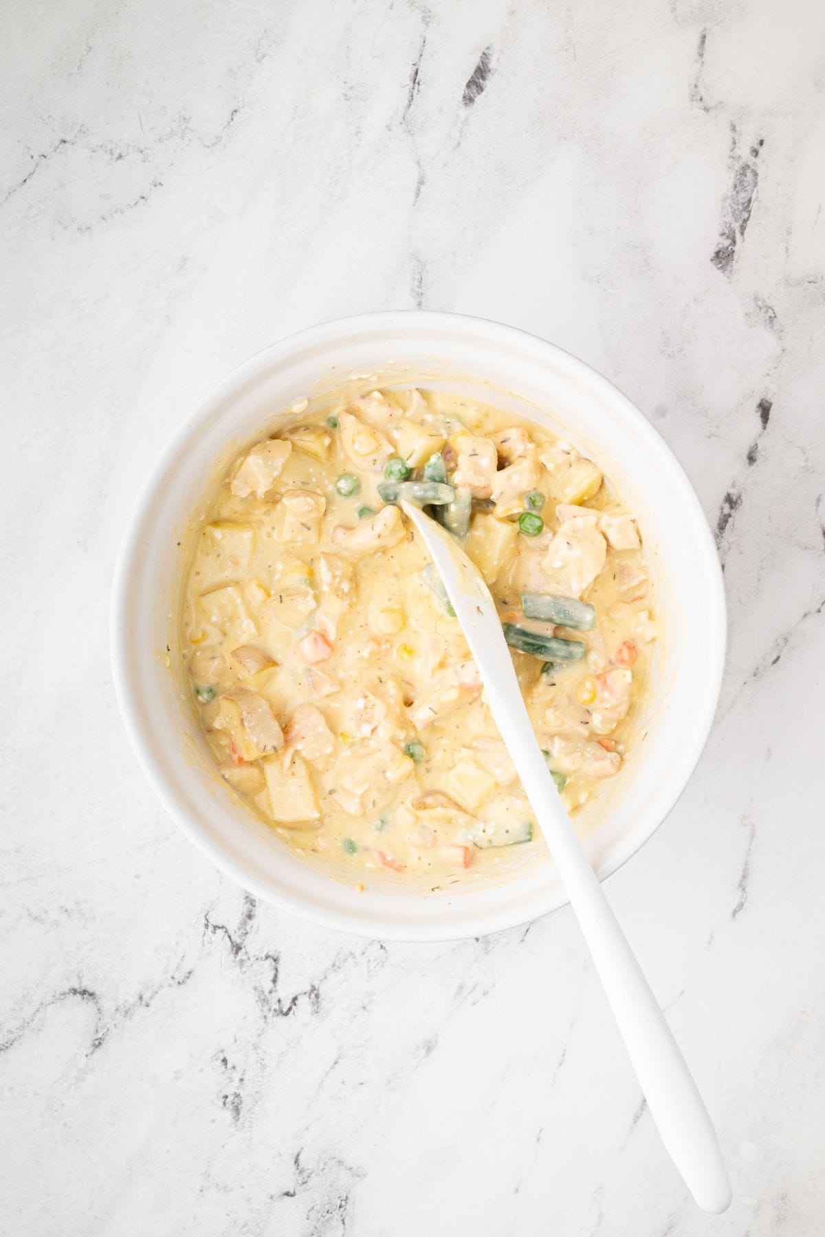 A bowl filled with a creamy chicken, potato, and vegetable mixture, stirred with a white spoon.
