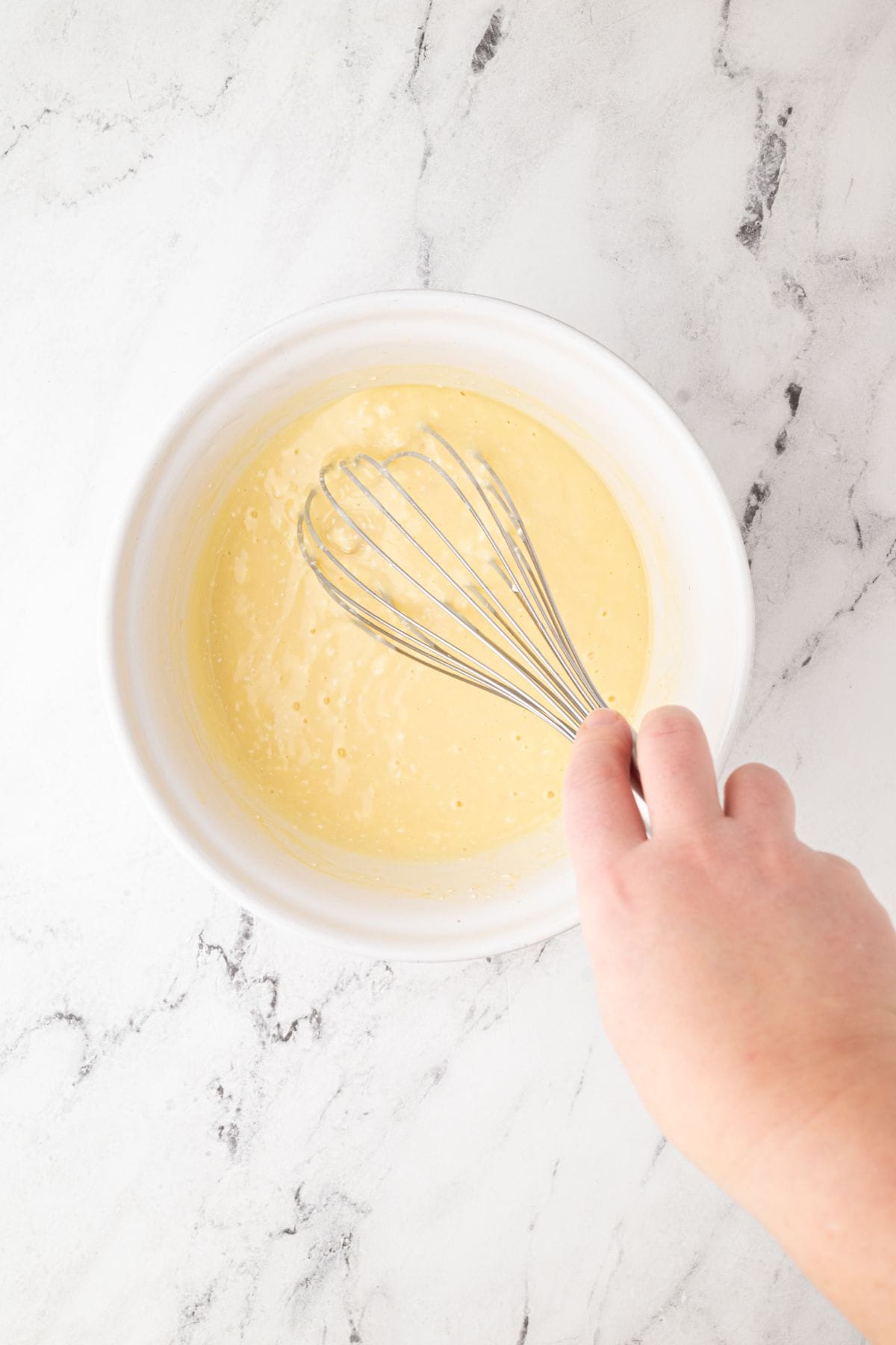 A bowl with a yellow batter being whisked by hand.