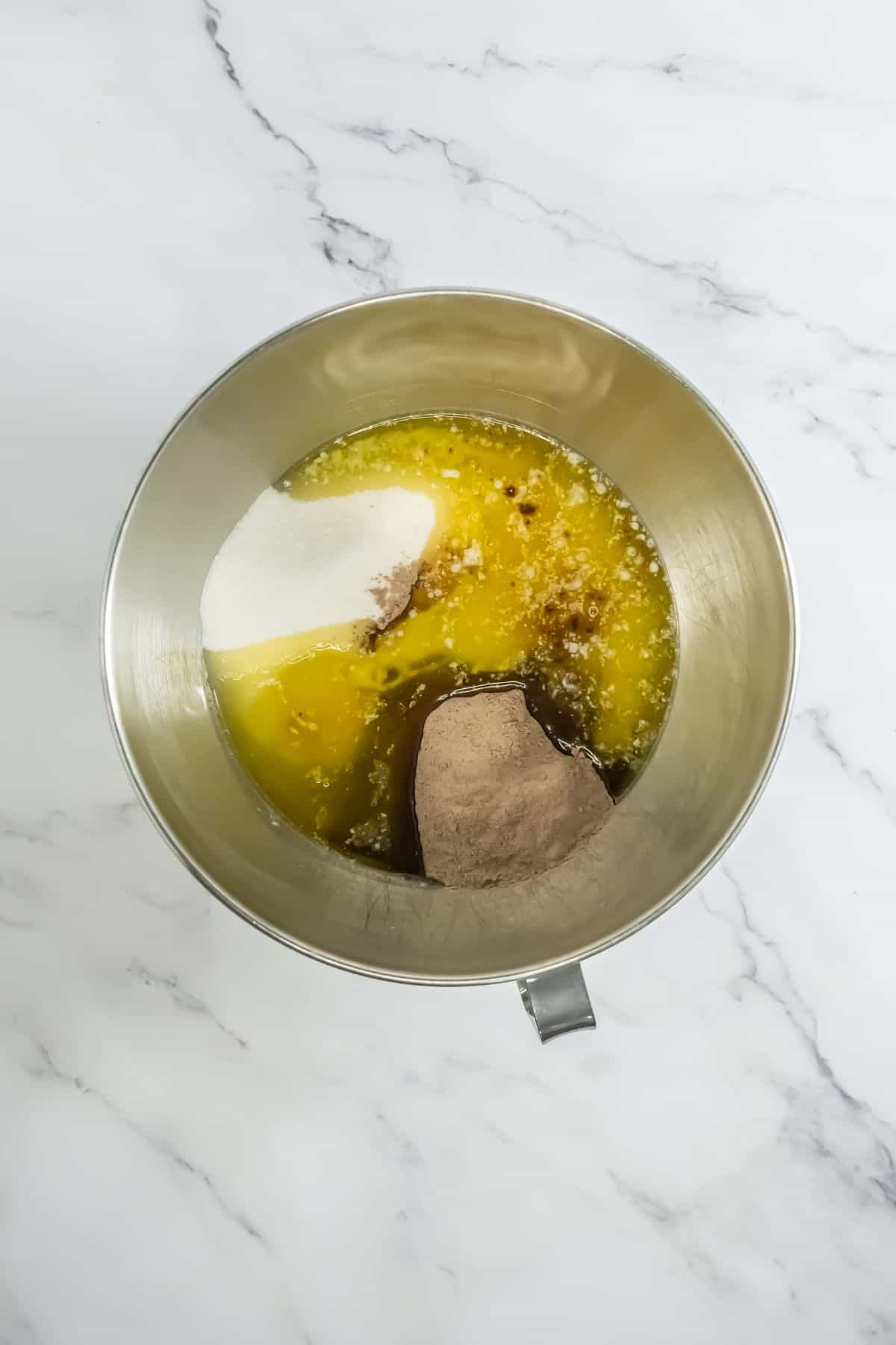 A mixing bowl with partially combined ingredients for the chocolate pudding cake. Visible ingredients include sugar, melted butter, cocoa powder, and pudding mix