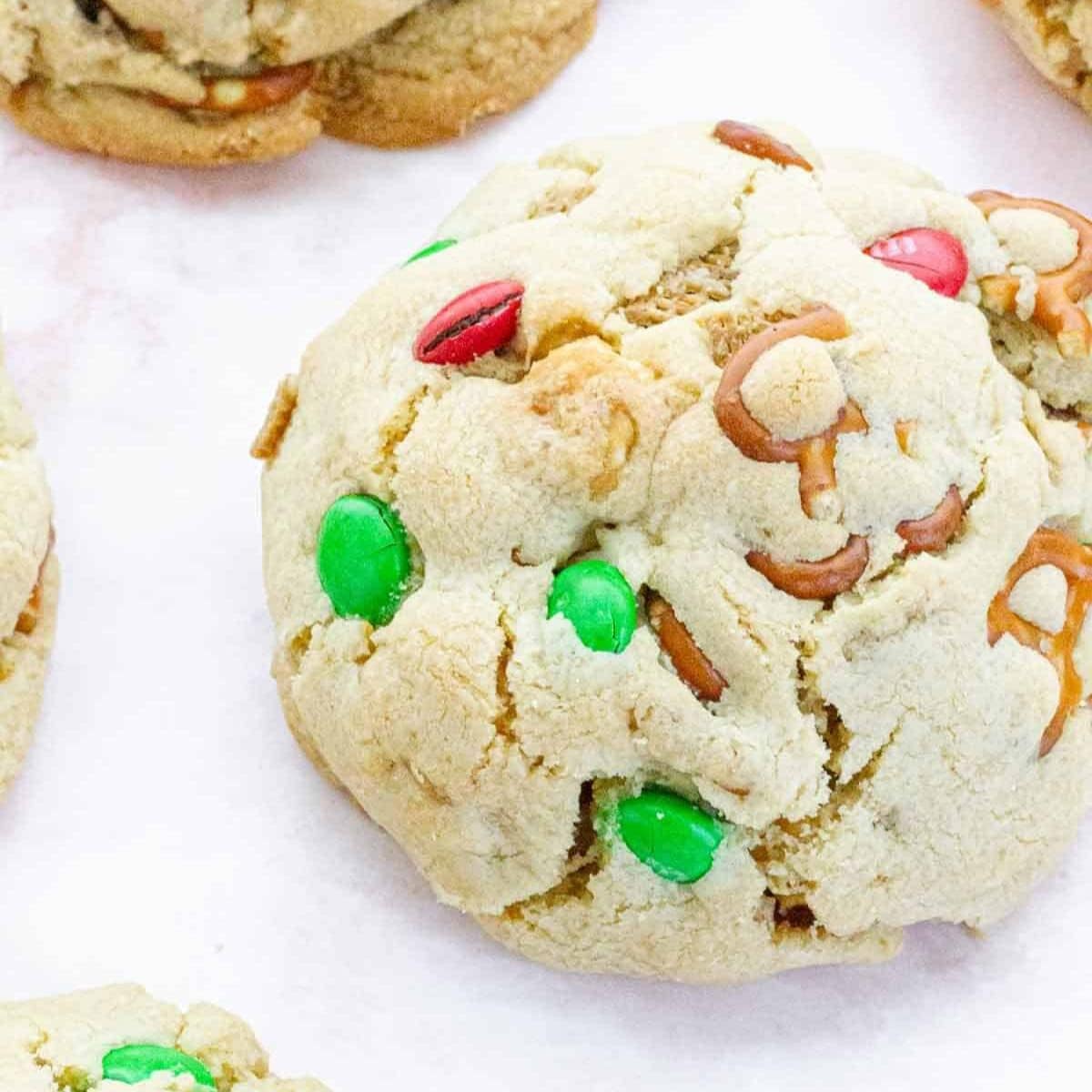 A close-up of a Christmas Chex Mix cookie, showing its golden, slightly cracked surface with pretzels, M&Ms, and cereal pieces embedded in the dough.