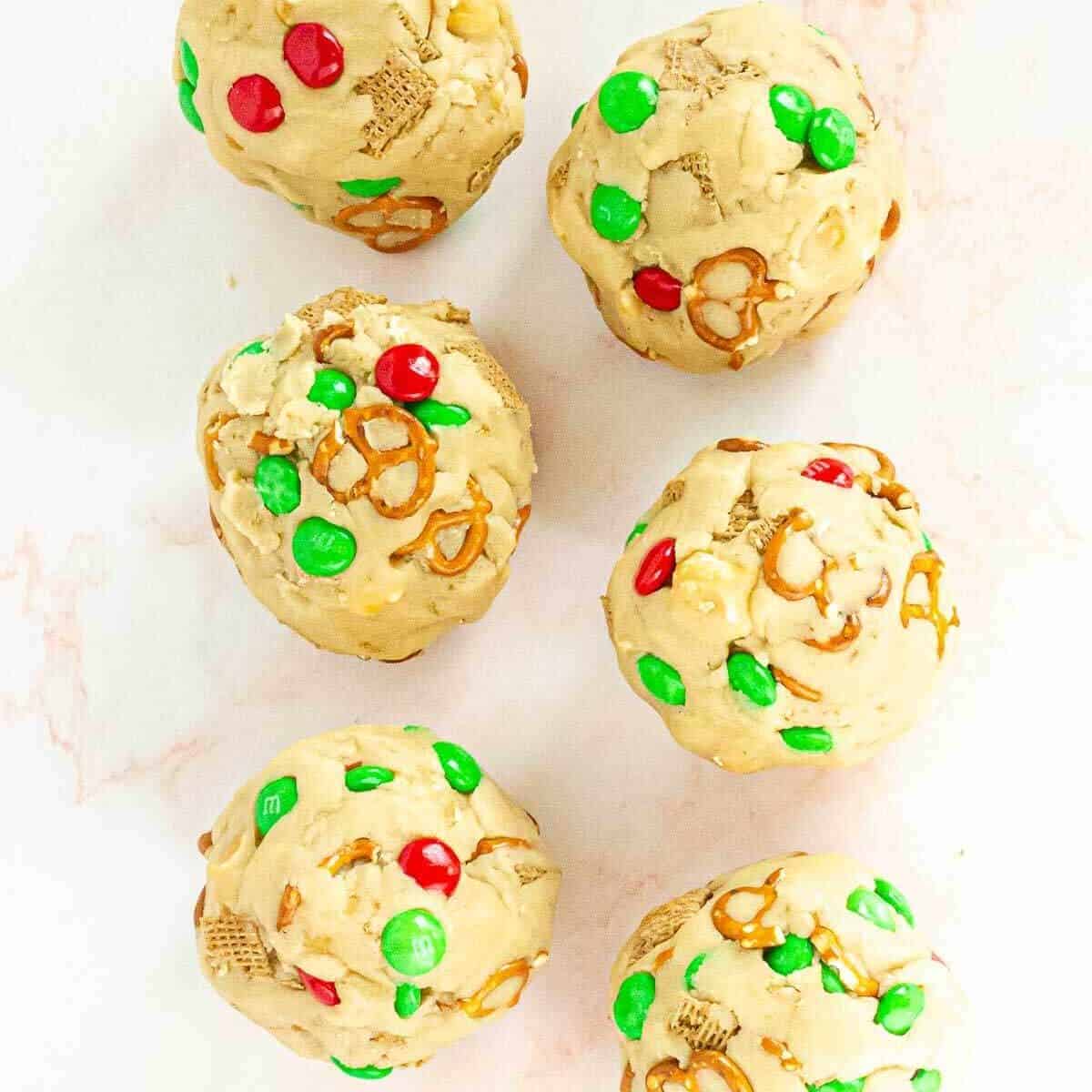 Six large, round cookie dough balls with pretzels, M&Ms, and cereal pieces.