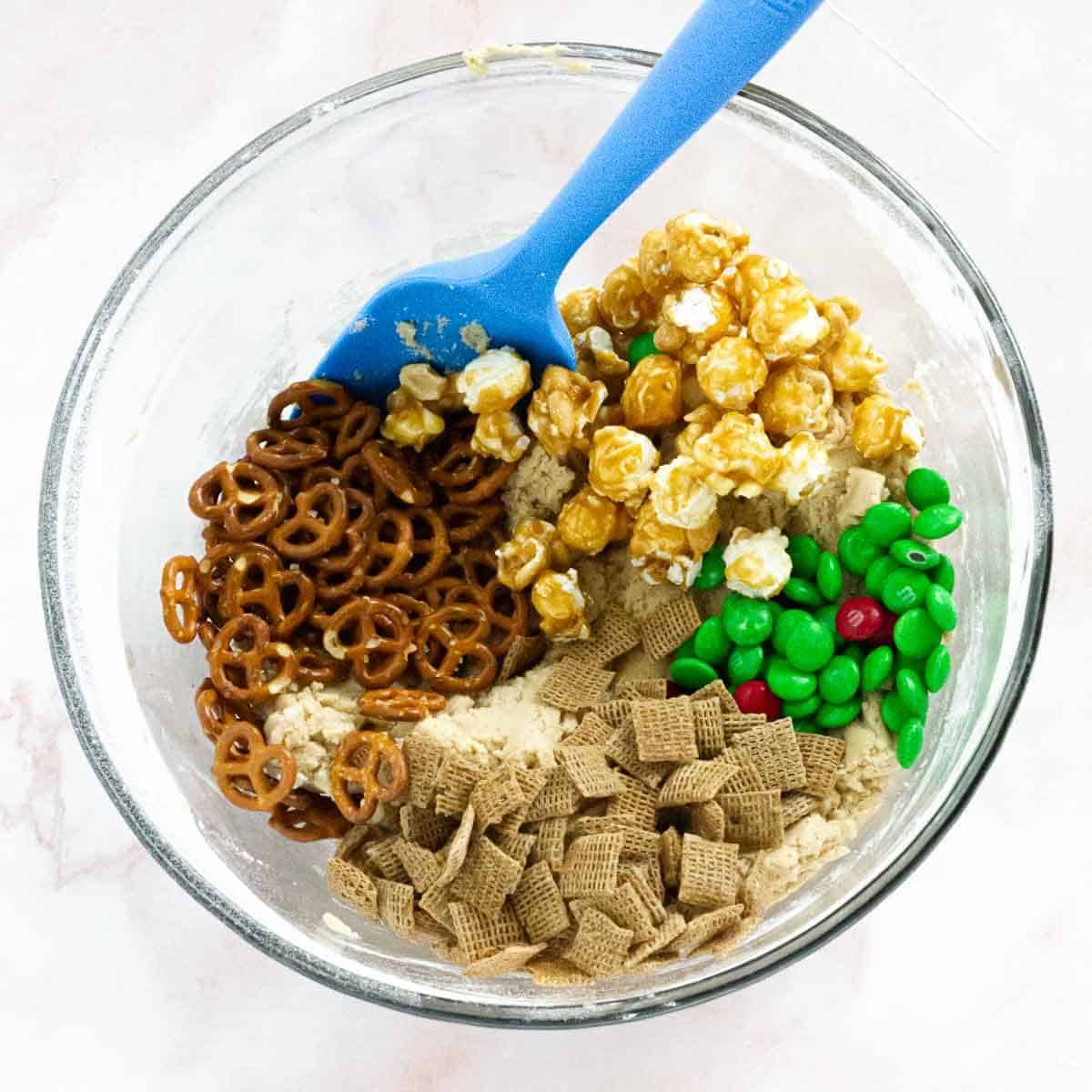 A bowl of dough with pretzels, caramel popcorn, Chex cereal, and red and green M&Ms before mixing.