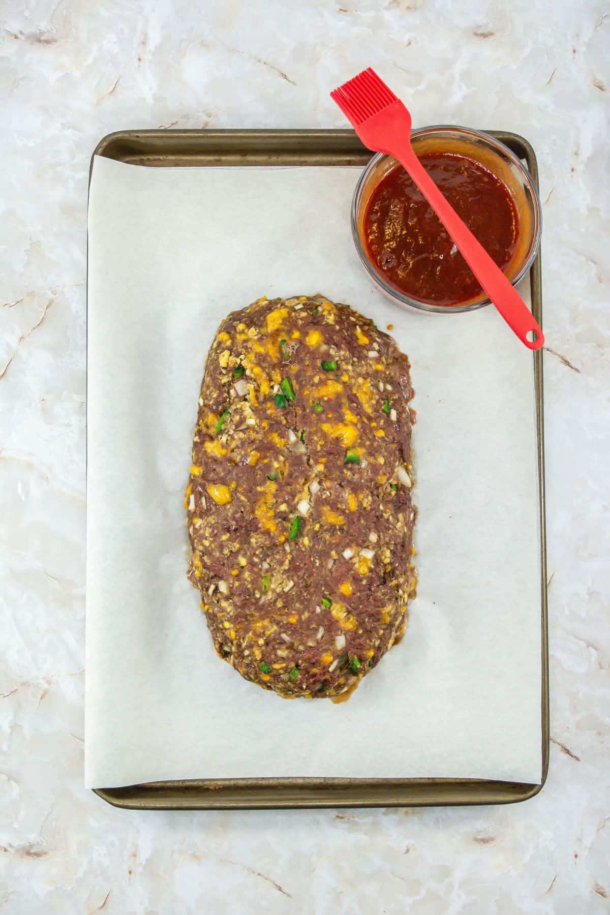a parchment-lined baking sheet with a half-baked meatloaf mixture and a small bowl of glaze next to it, with a basting brush resting on the bowl