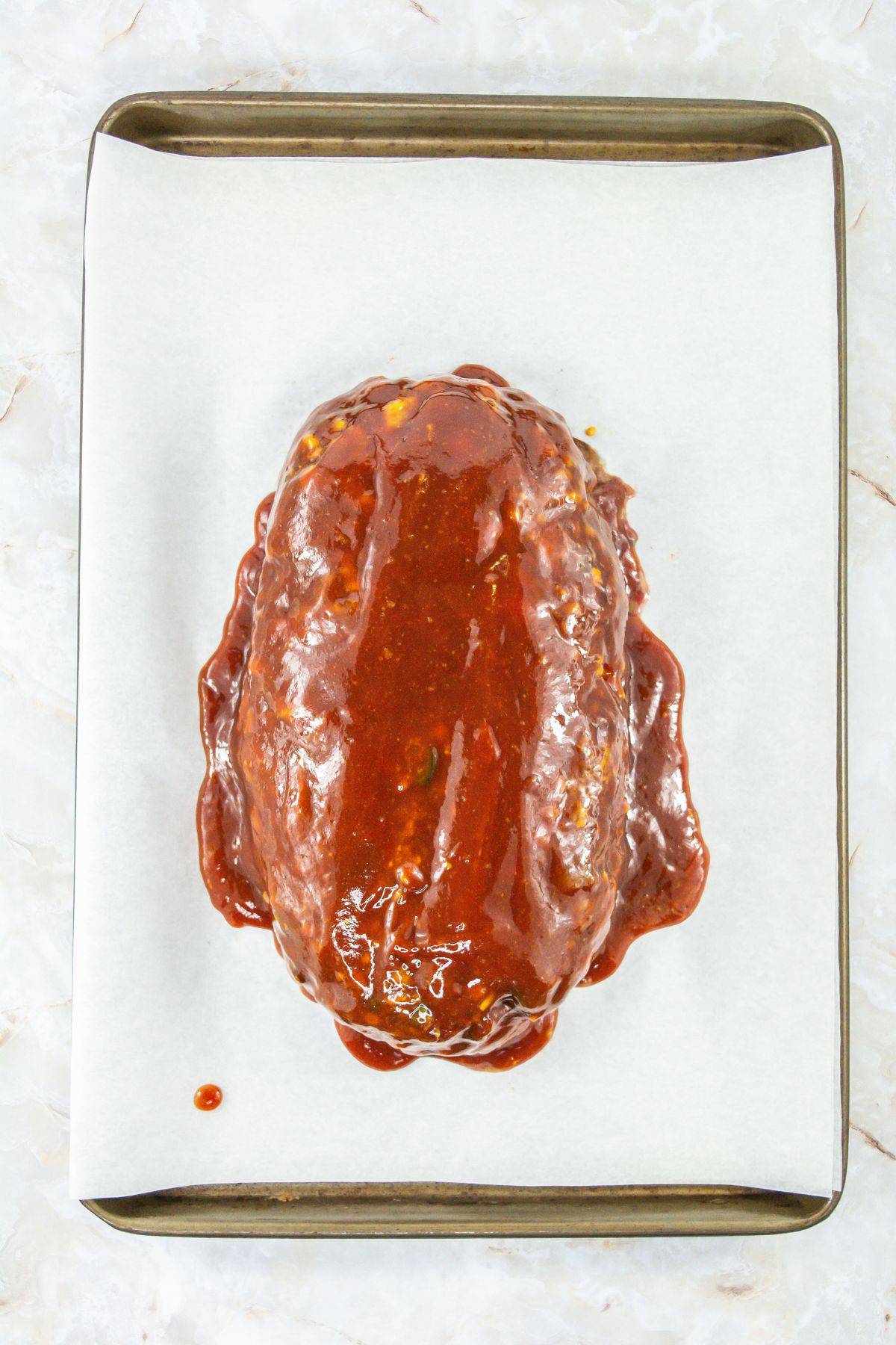 half-baked meatloaf mixture coated with a thick layer of glaze on a parchment-lined baking sheet