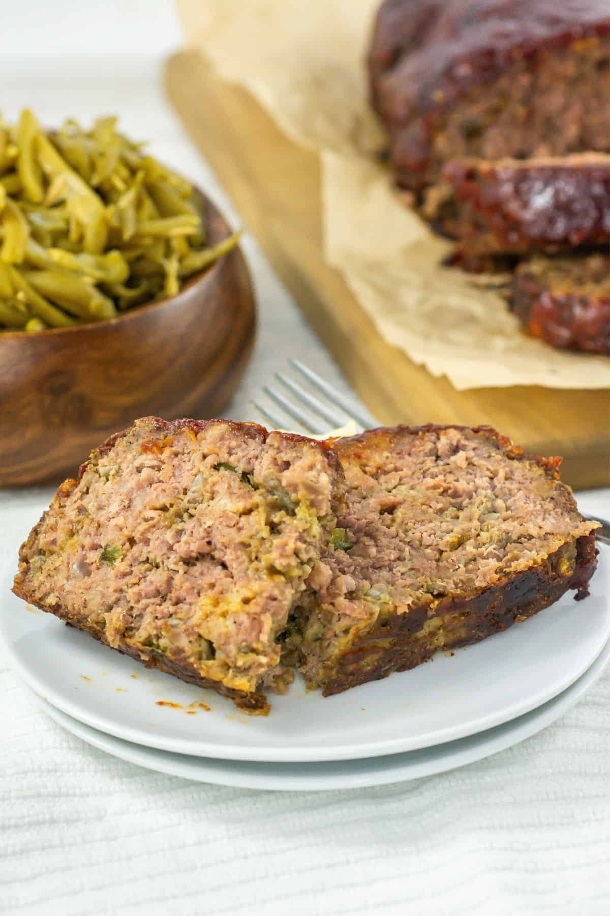 a sliced meatloaf on a white plate, and a bowl of cooked green beans and copycat cracker barrel meatloaf on parchment paper on a wooden cutting board in the background