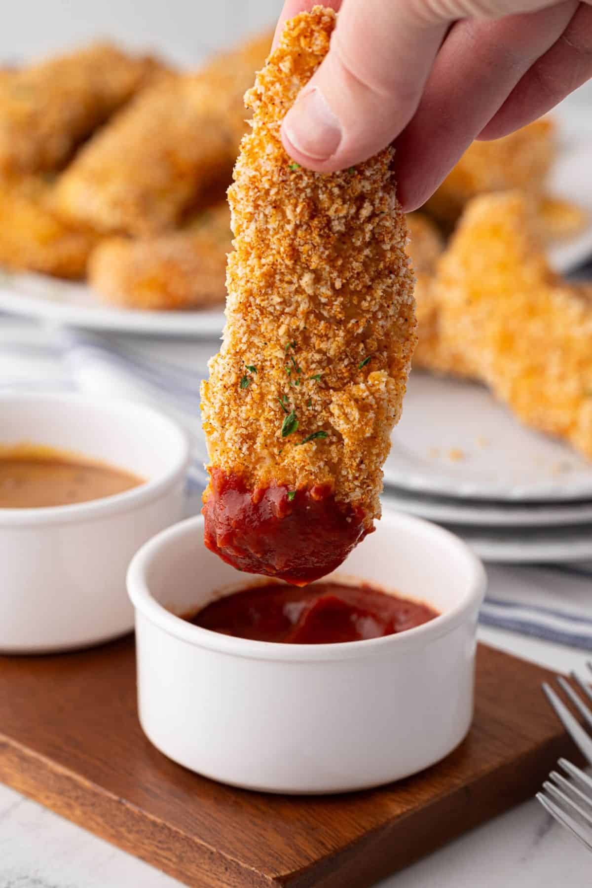 A golden-brown tender being dipped into ketchup.