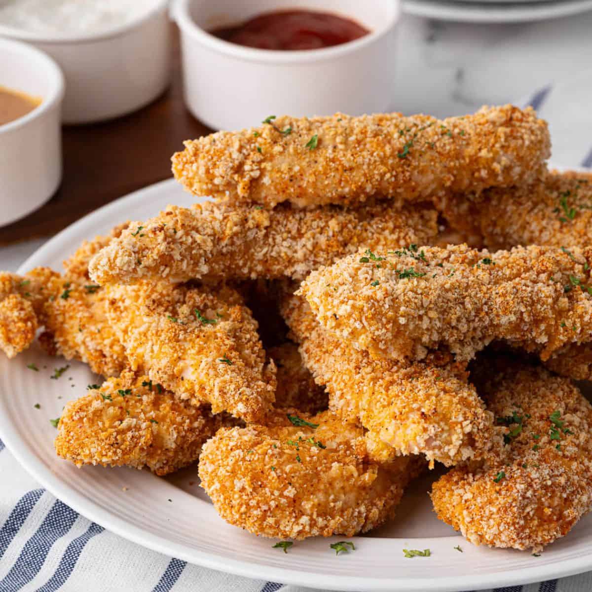 A pile of crispy baked chicken tenders served on a plate, garnished with parsley, alongside dipping sauces.