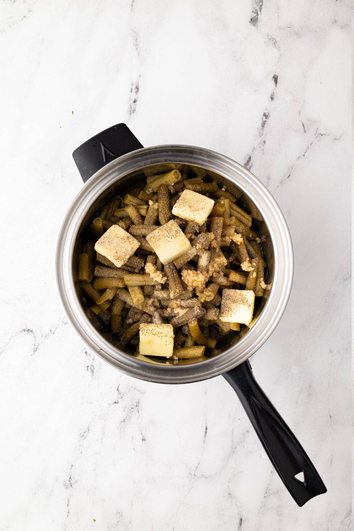 Canned green beans in a saucepan with butter cubes, minced garlic, and seasonings before cooking.