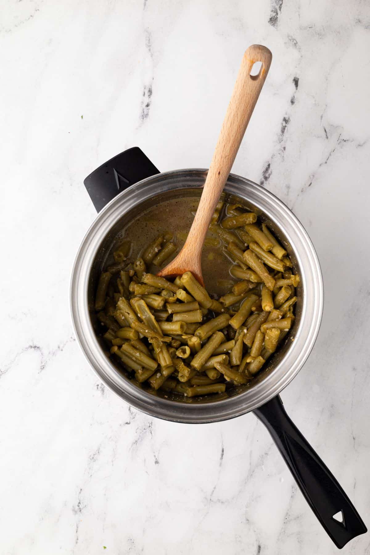 Cooked green beans in a saucepan, stirred with a wooden spoon, showing a rich, savory sauce.