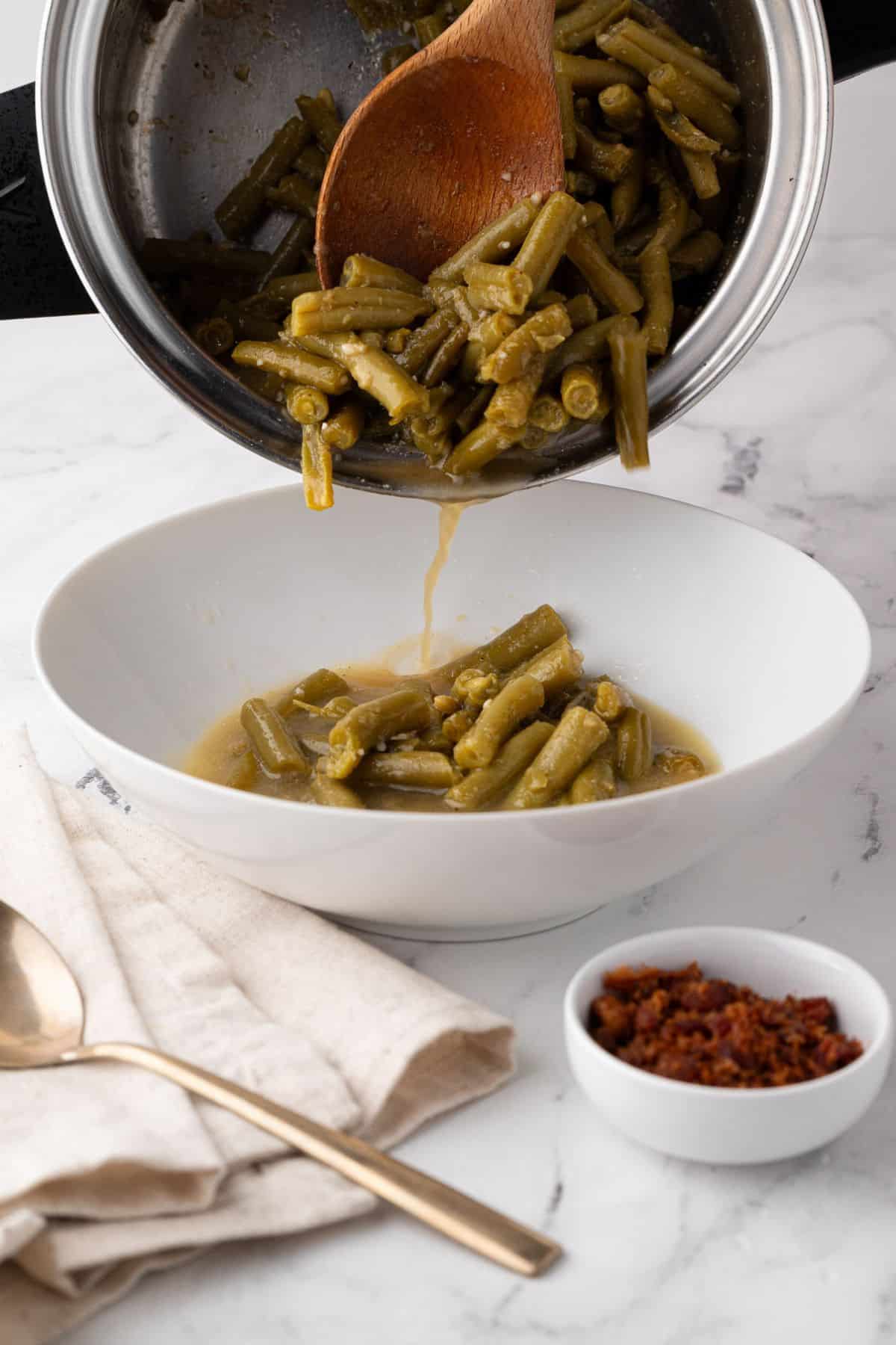 Green beans being poured from a saucepan into a serving bowl, with crispy bacon bits in a dish beside it.