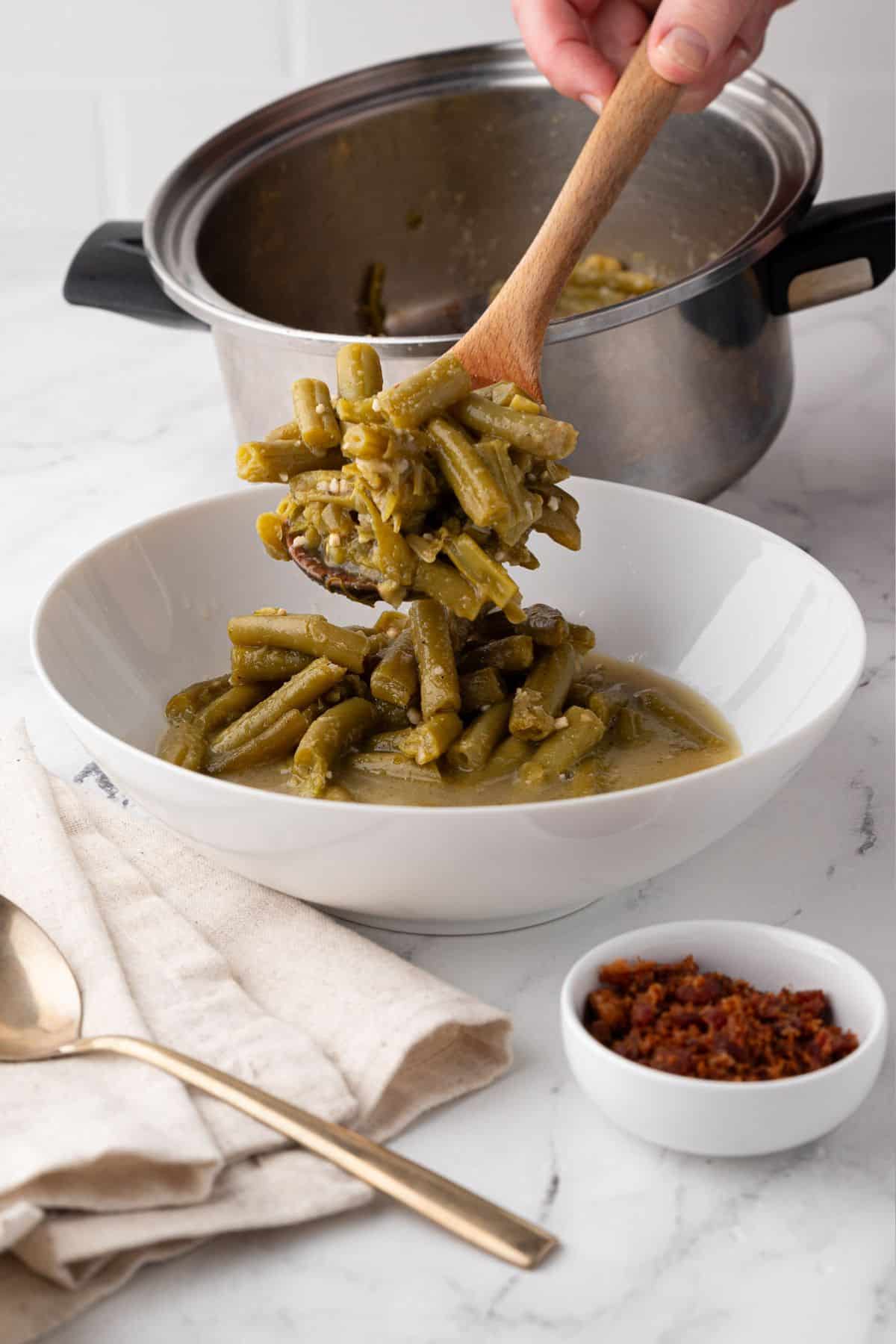 Cooked Canned green beans being served from a saucepan into a bowl, with crispy bacon bits in a small dish beside it.