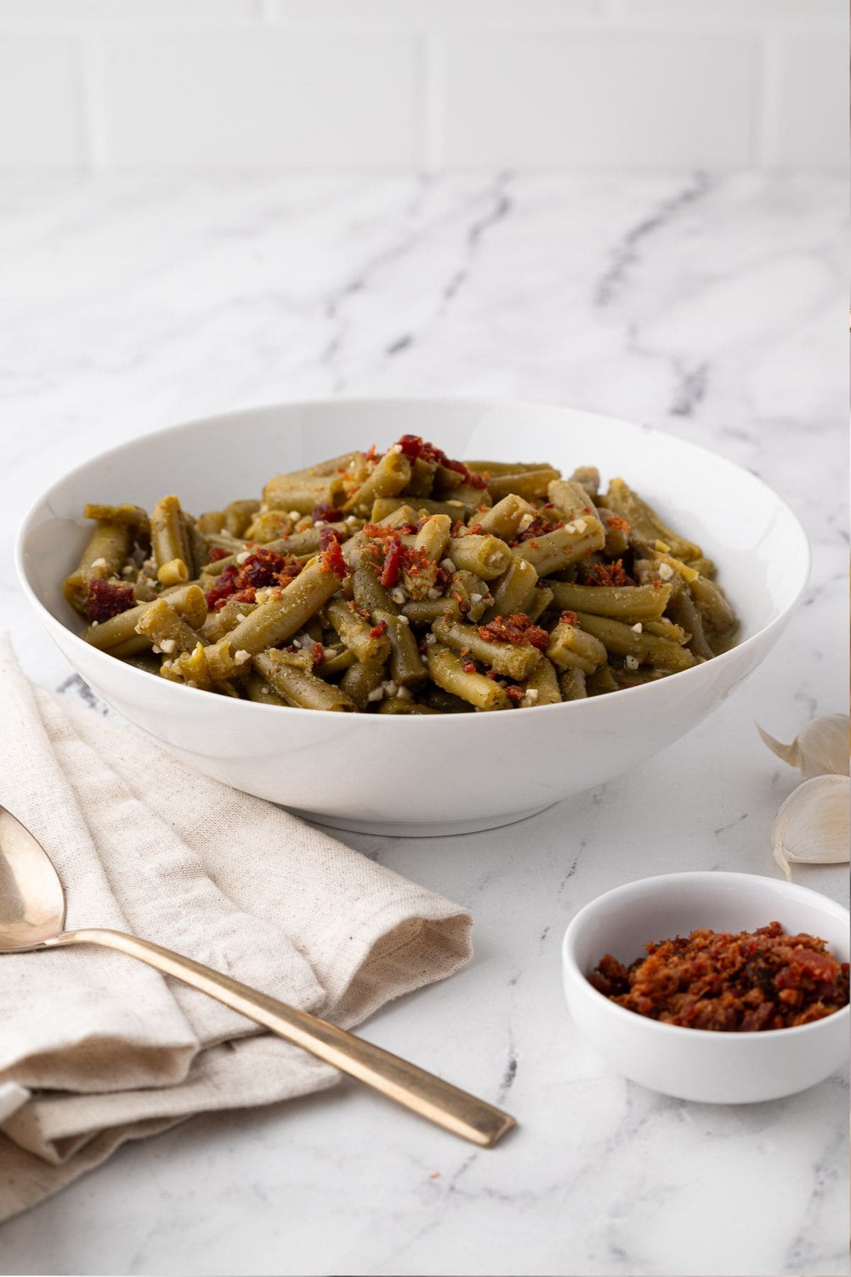 Green beans being poured from a saucepan into a serving bowl, with crispy bacon bits in a dish beside it.