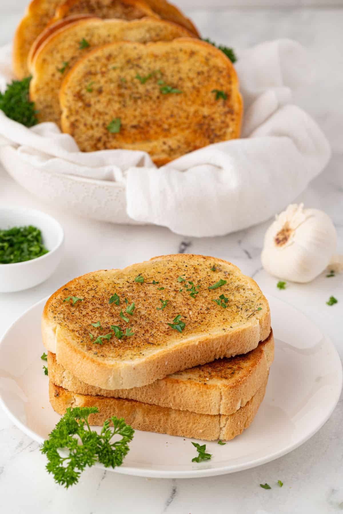 Stacked garlic toast on a white plate, garnished with fresh parsley and surrounded by garlic and herbs.