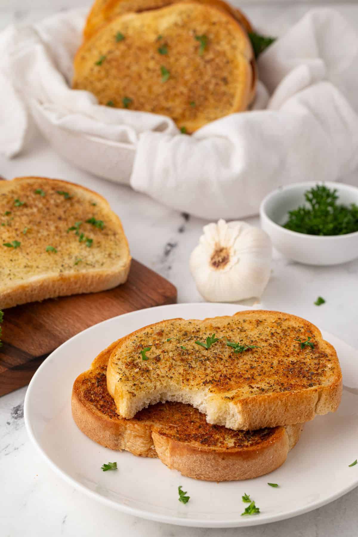 Toasted garlic bread slices on a plate with a bite taken out, garnished with parsley and served with garlic in the background.