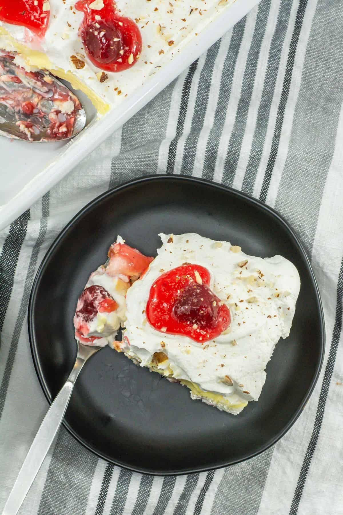 A slice of dessert on a black plate with a forkful taken out, showing layers of cake, cream, and cherries.