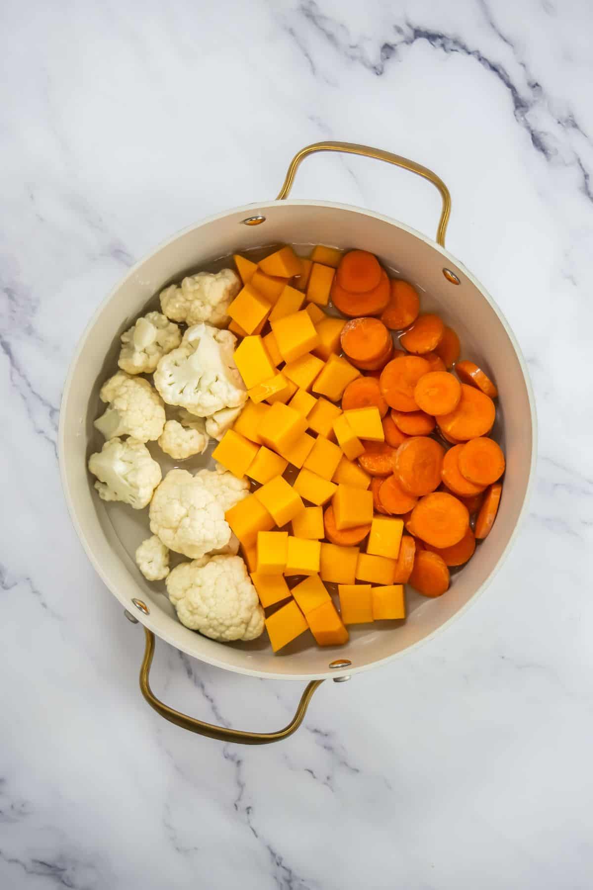 Overhead view of a pot with raw chopped carrots, cauliflower, and butternut squash prepared for cooking.